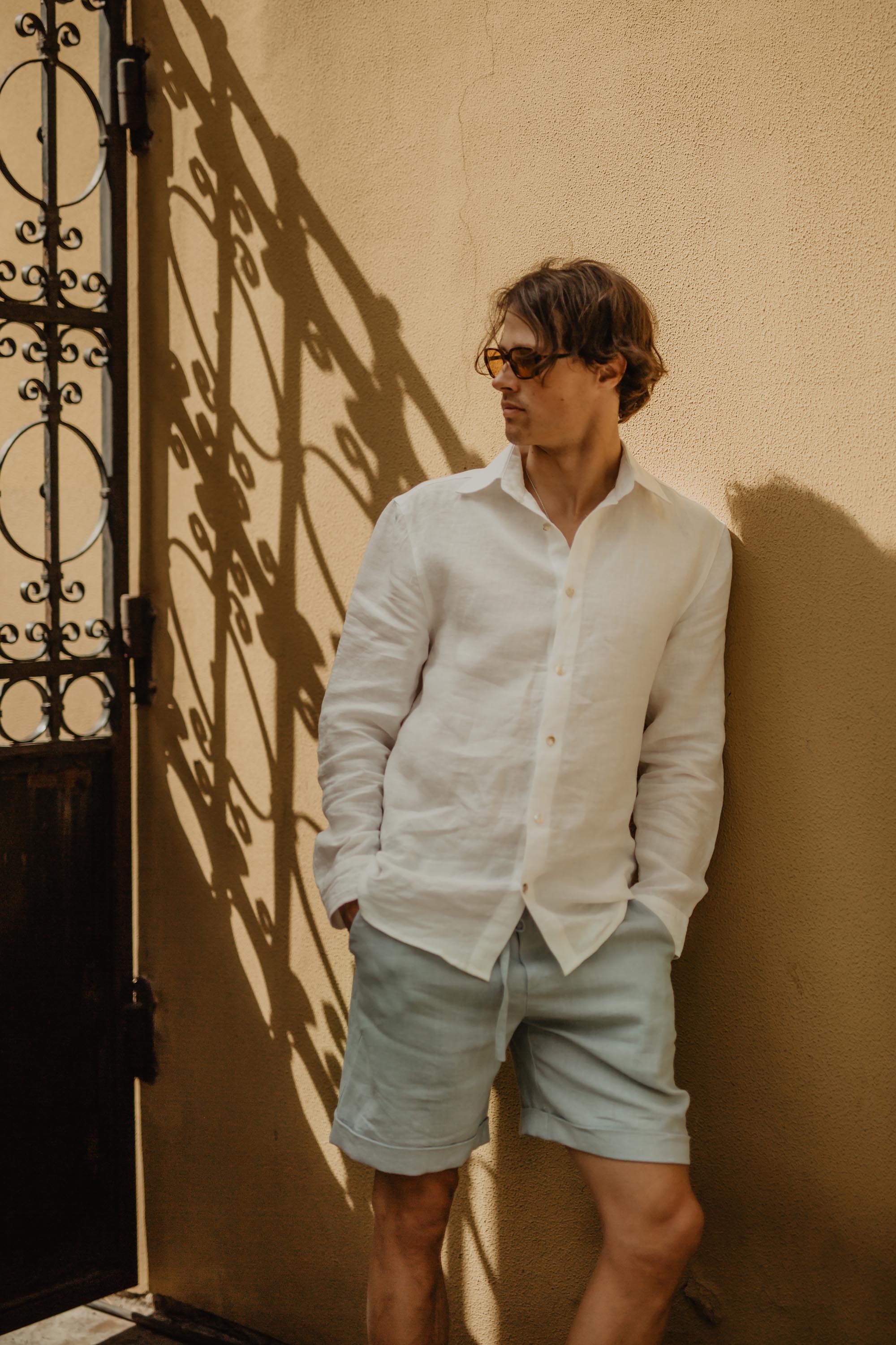 Man Leaning On A Wall Wearing Sage Green Linen Shorts And White Shirt