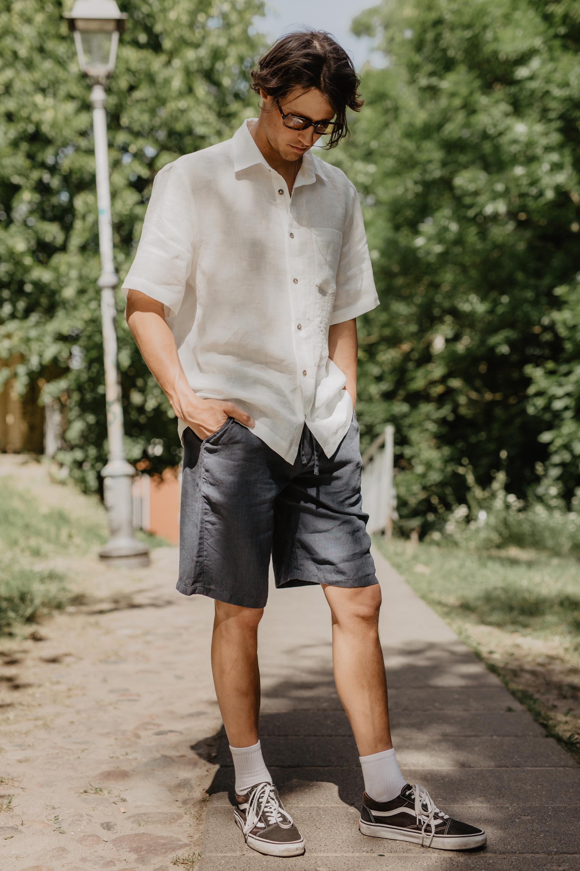Man Looking Down At His Shoes Wearing White Oversized Linen Shirt And Black Linen Shorts