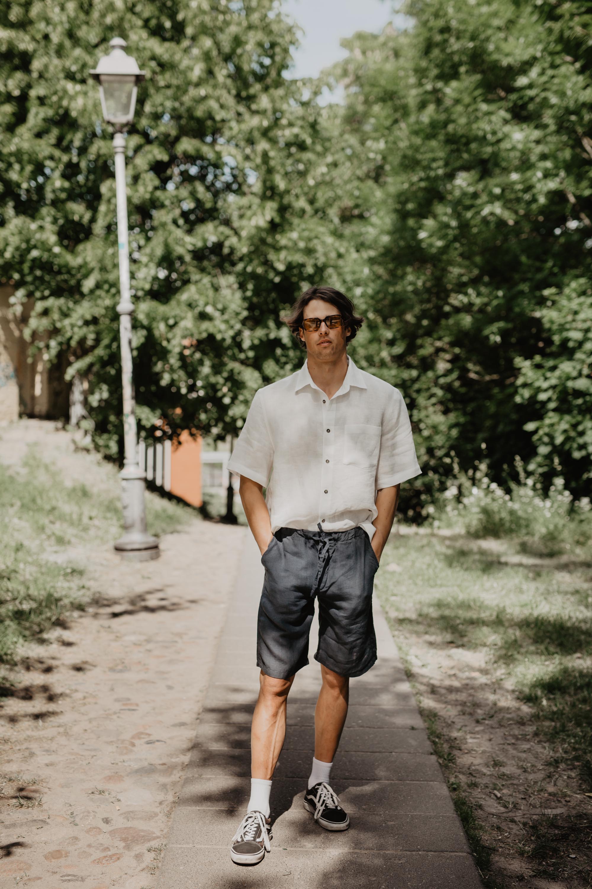 Man With Hands In Pockets Standing On Walkway Wearing White Oversized Linen Shirt and Black Linen Shorts