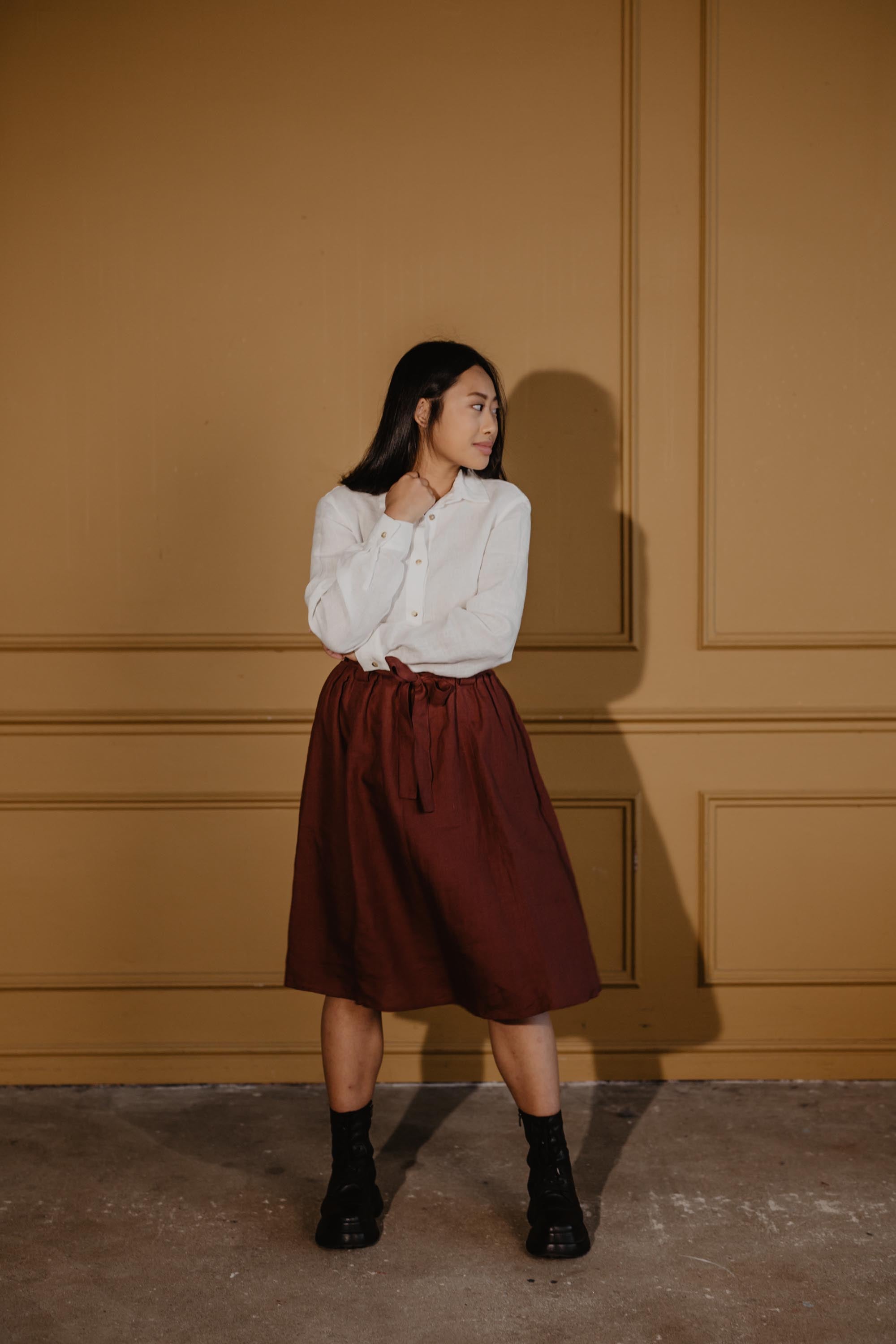 Woman Looking to The side Wearing A Terracotta Bergen Mid-Length Linen Skirt and White Linen Shirt By Amourlinen