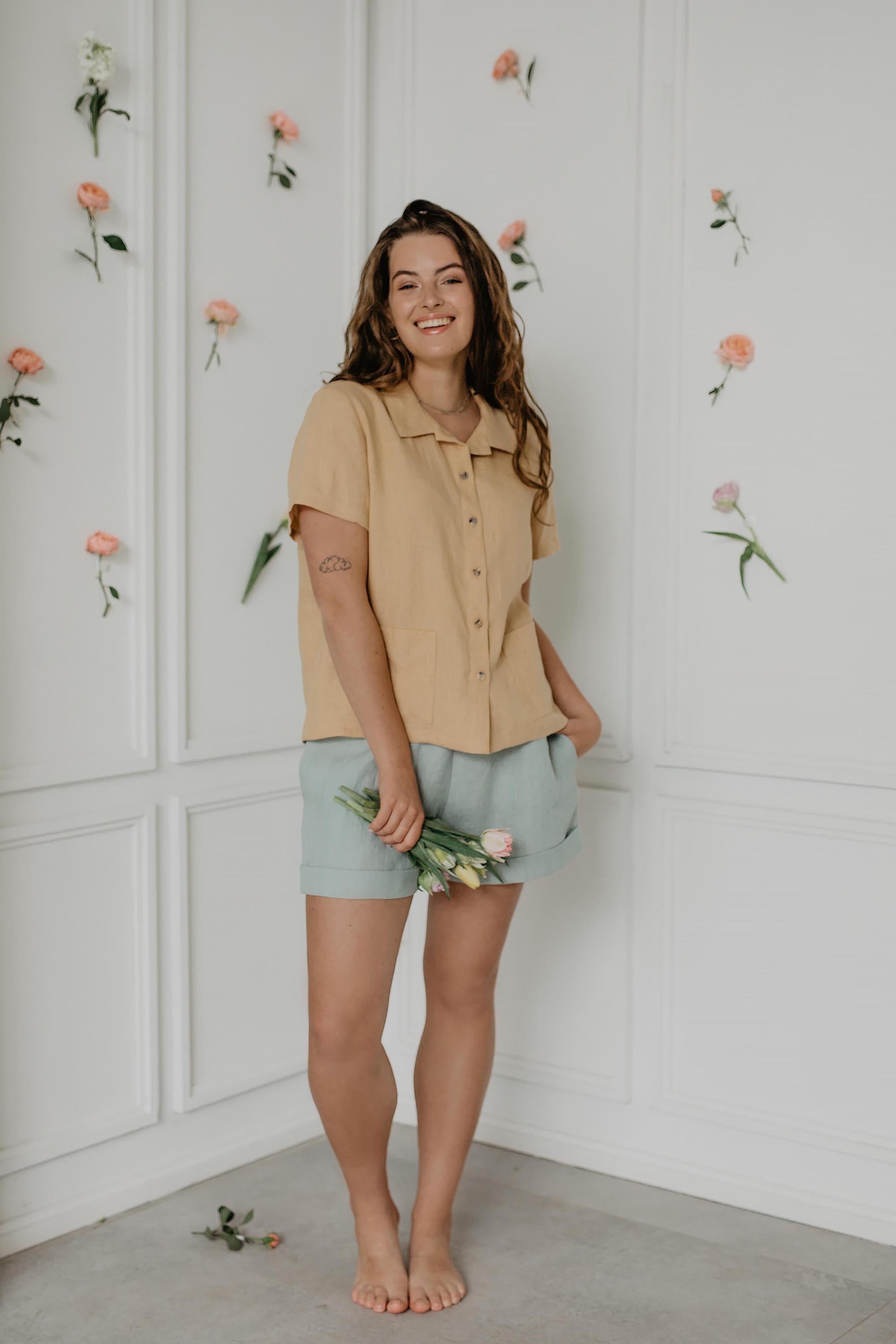Woman Smiling In White Room With Flowers Wearing Mustard Summer Linen Shirt and Sage Green Shorts