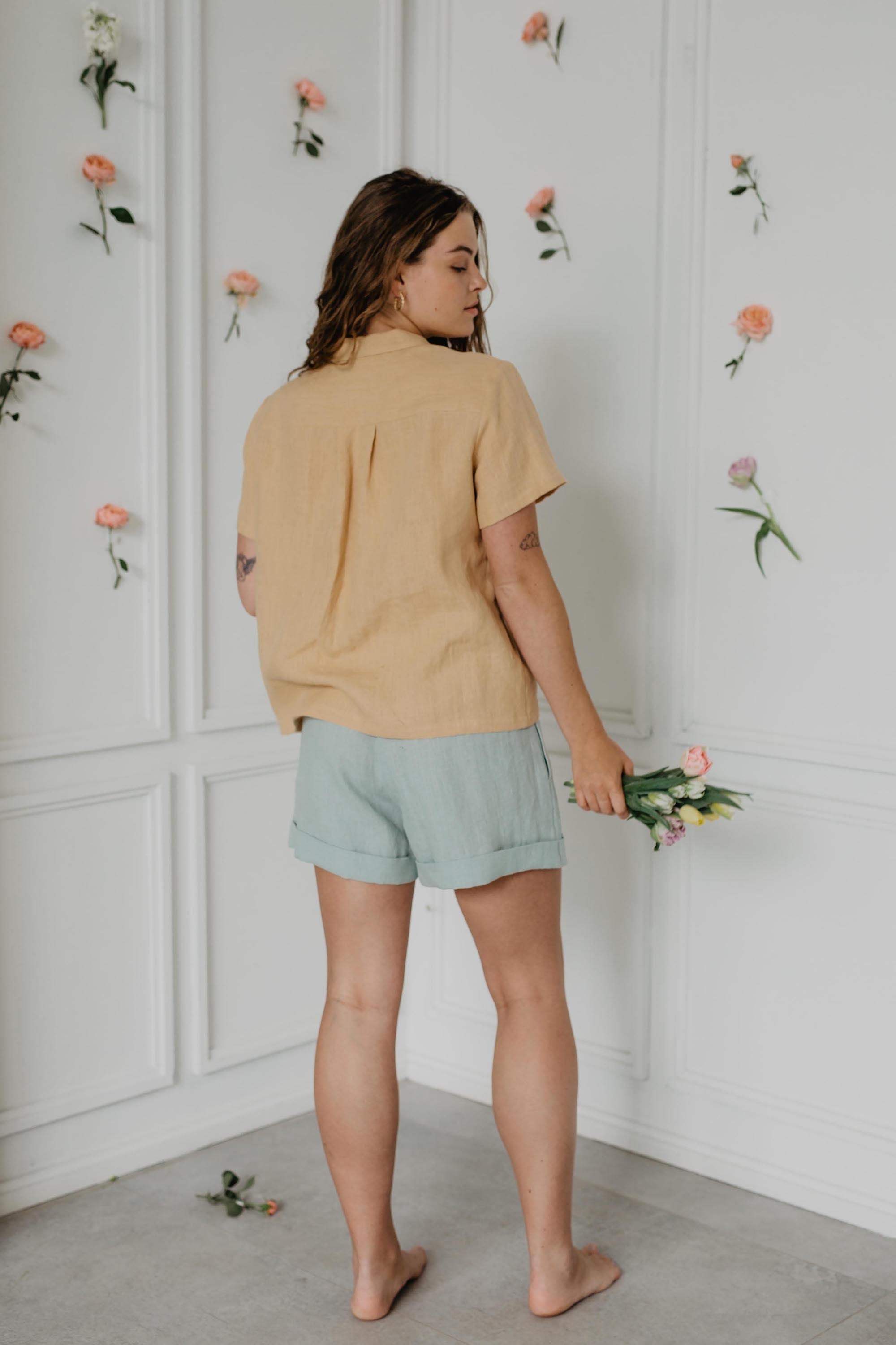 Back Of Woman Wearing Sage Green Linen Shorts and a Short Sleeve Mustard Shirt In a Light Flowery Room