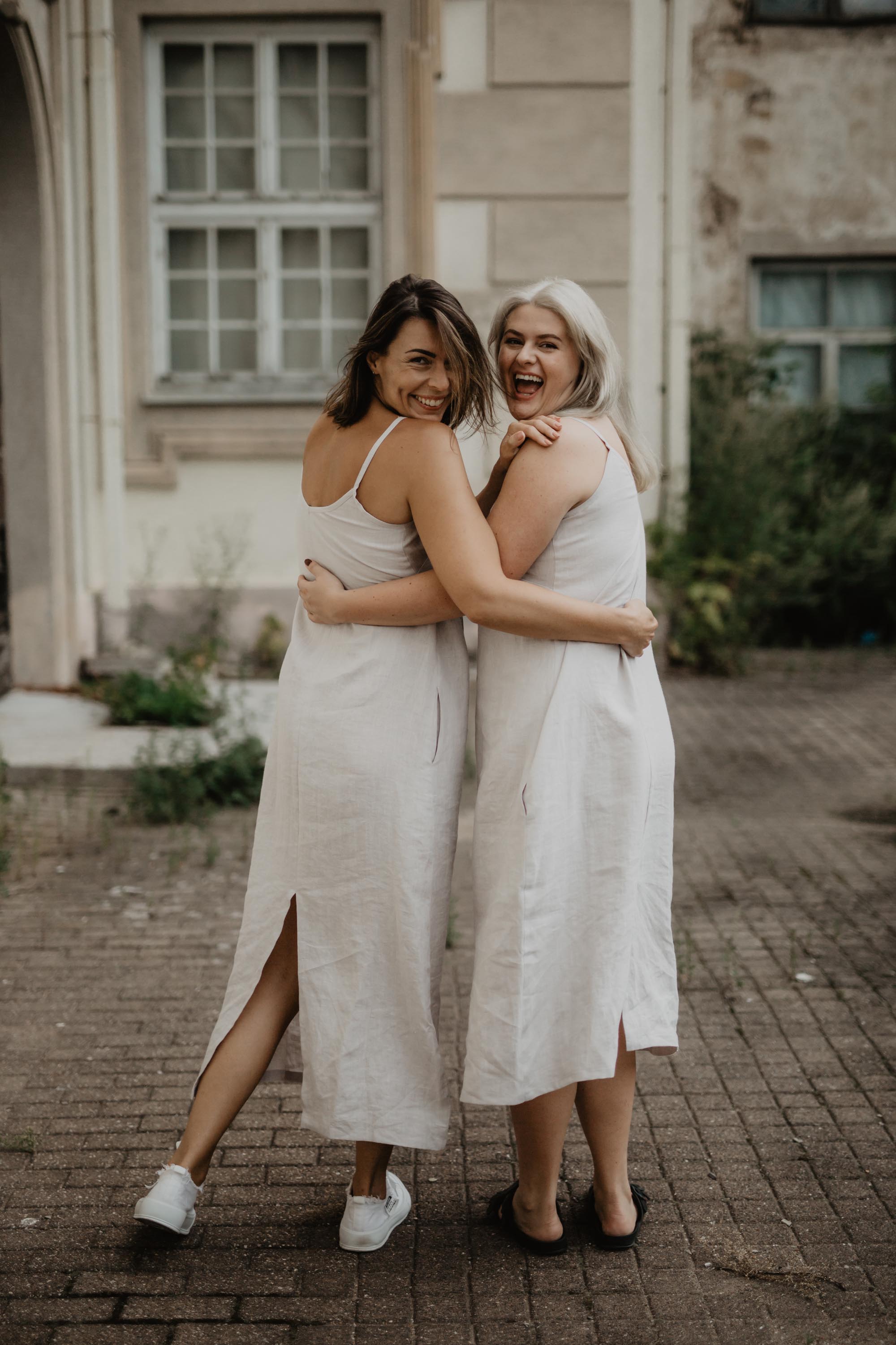 Two Women Laughing Wearing White Long Linen Desses BY AmourLinen In A Rustic Courtyard