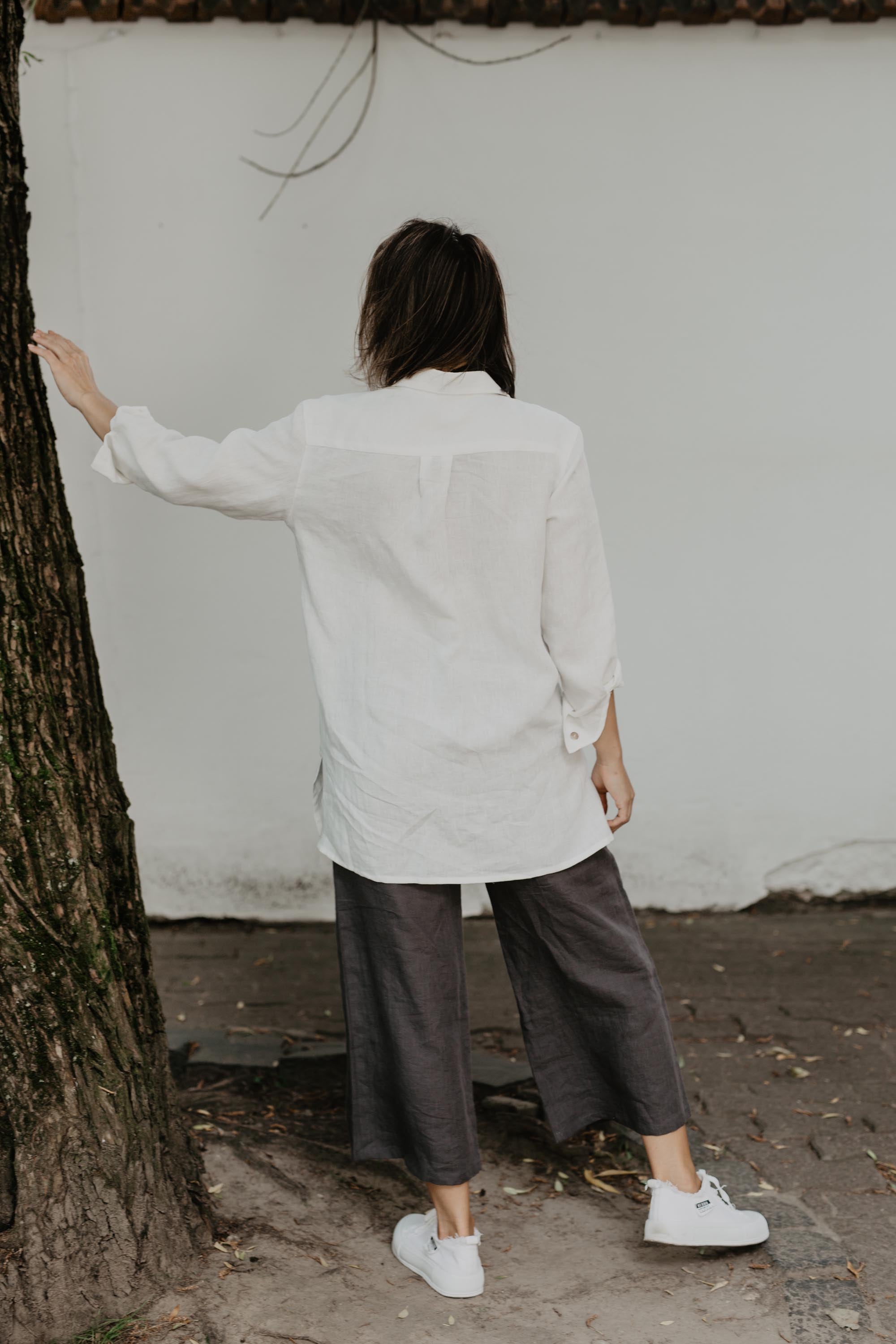 Woman Facing Back Wearing White Oversized Linen Shirt and Grey Pants Leaning Against a treaa