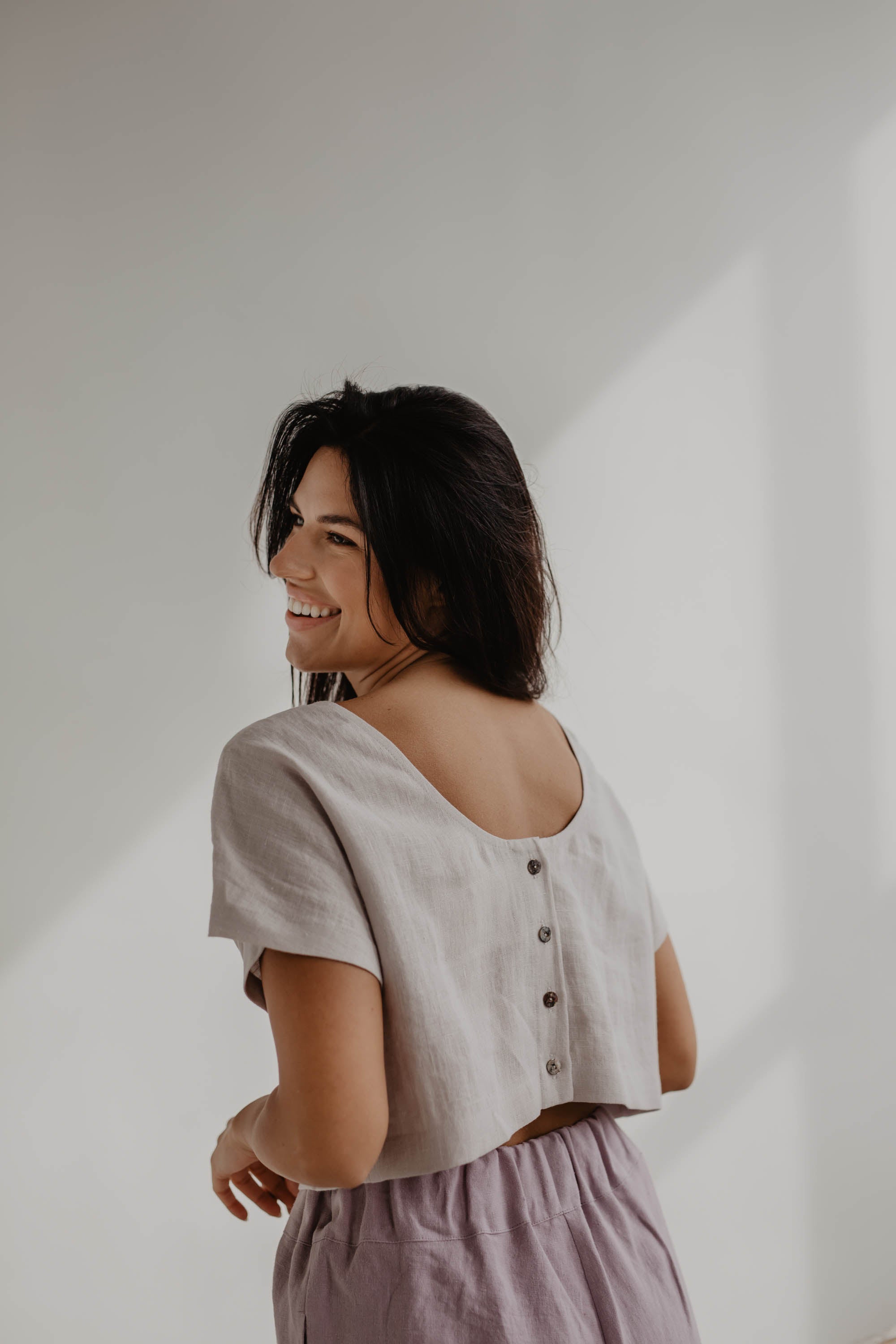 Woman Facing Back Wearing A Cream Color Linen Crop Top With Buttons