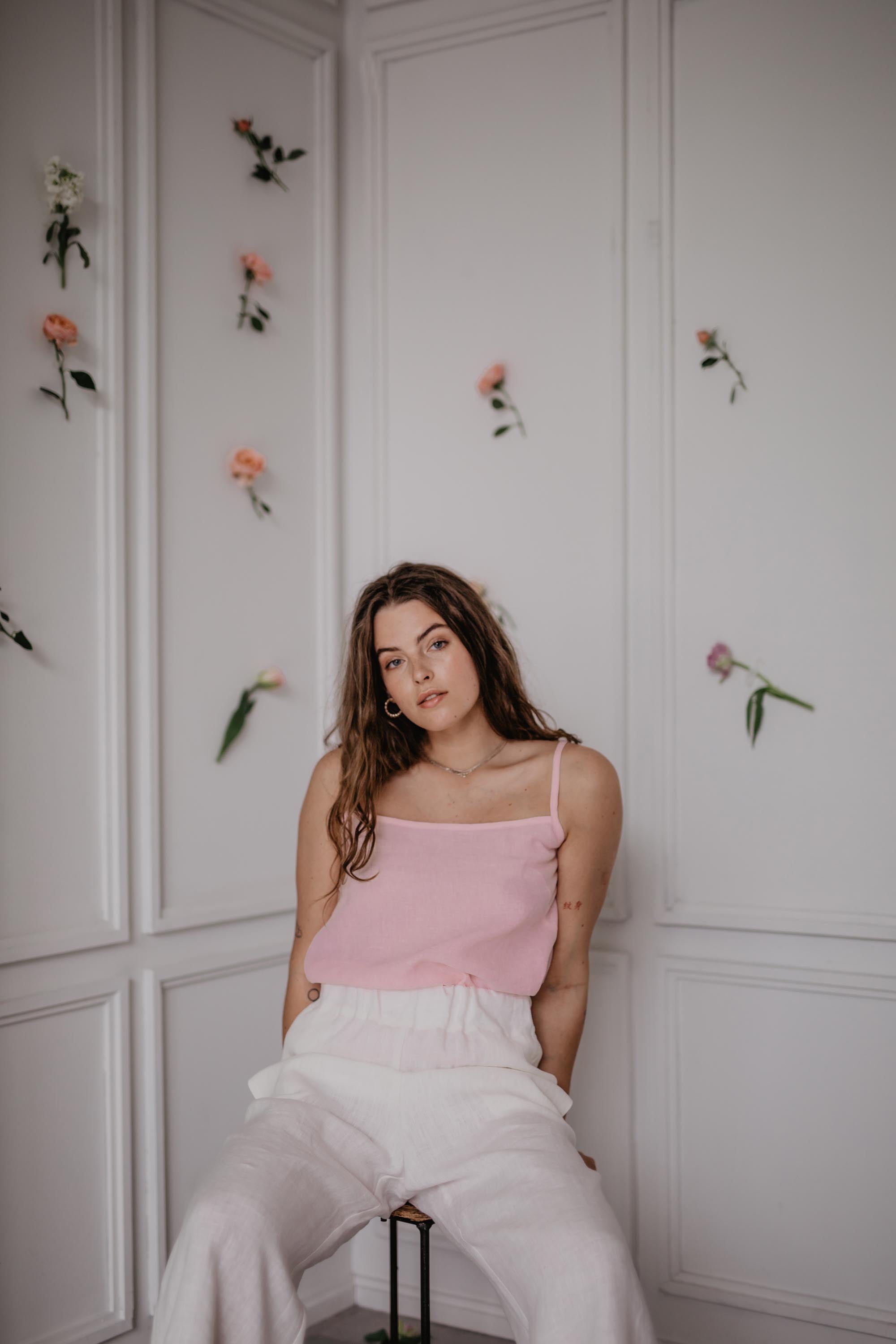 Woman Sitting In A Flowery Room Leaning Back Wearing A Pink Linen Top and Whitte Linen Pants By Amourlinen