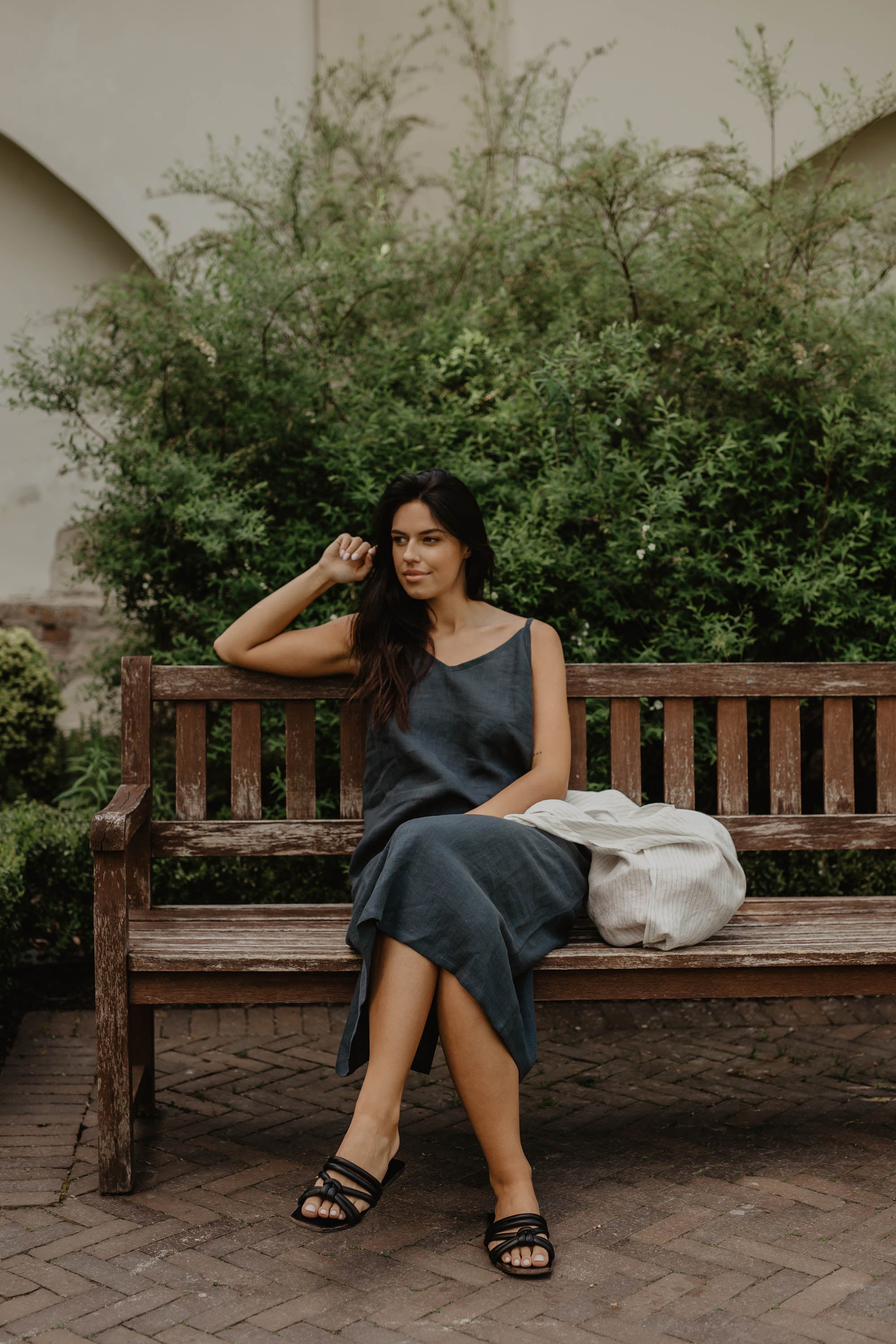 Woman Sitting On A Bench Wearing A Long Dark Linen Dress And A White Linen Tote Bag By AmourLinen