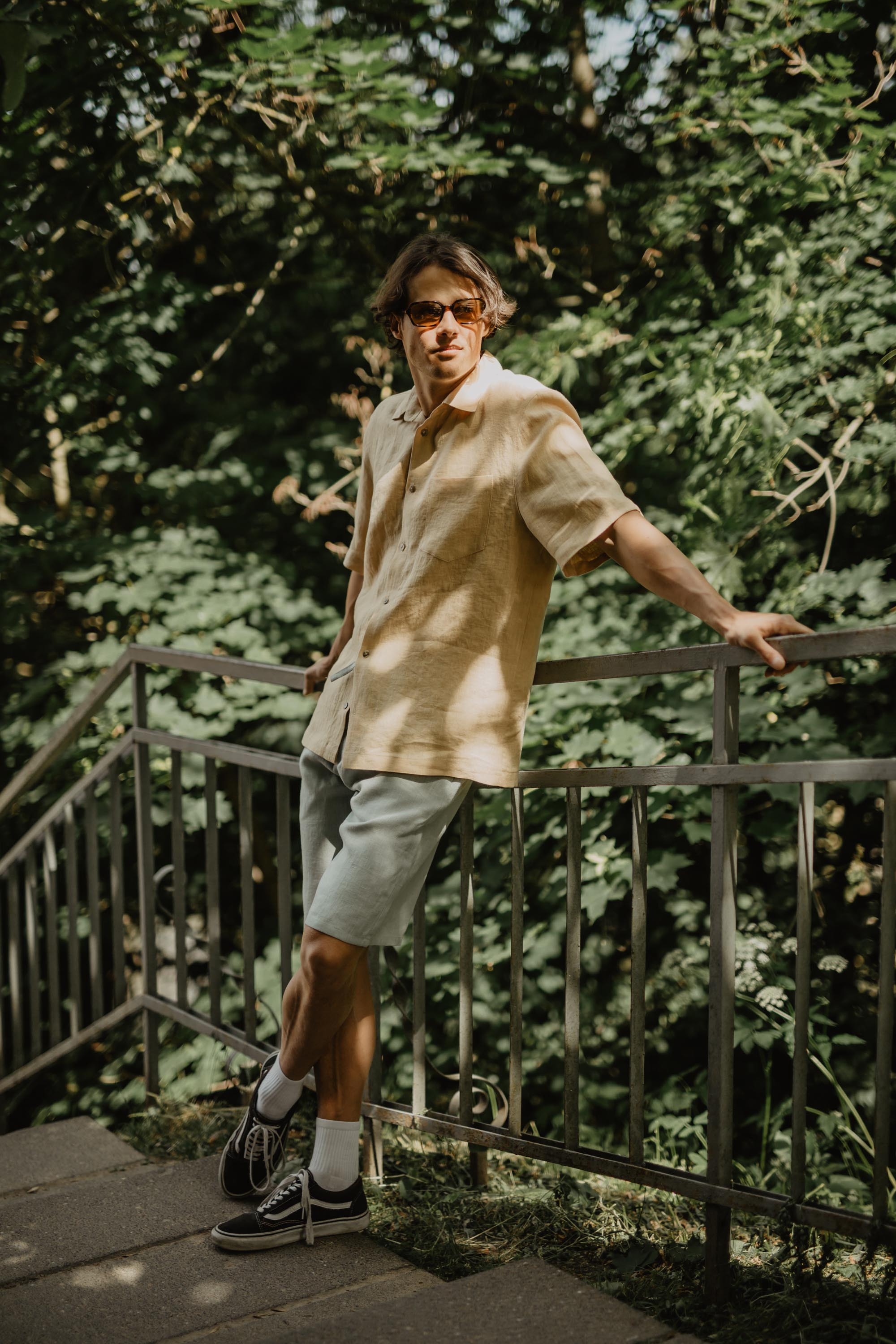 Man Leaning Against Railing Wearing Mustard Oversized Linen Shirt and Sage Green Linen Shorts