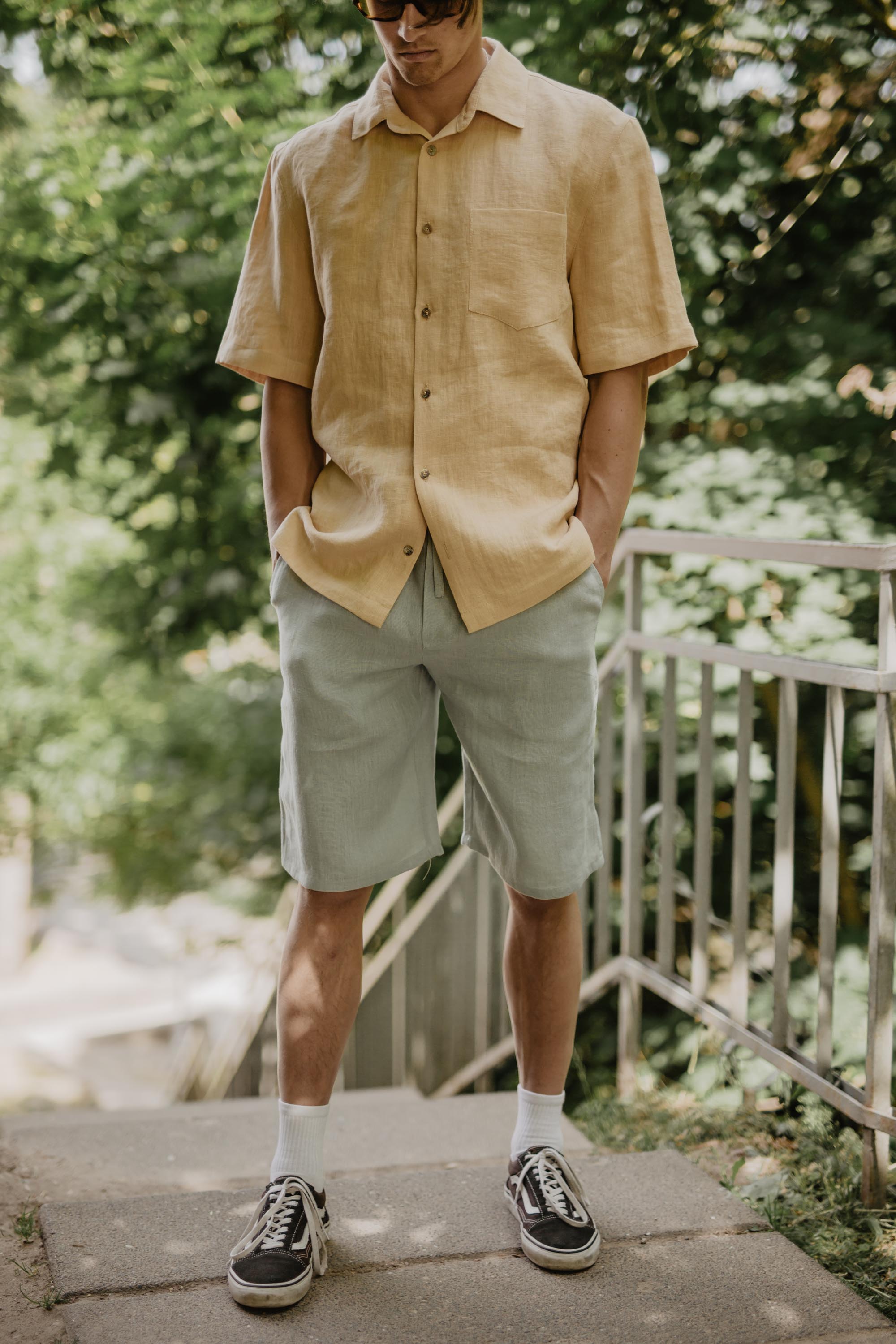 Man With Hands In Pockets Wearing Mustard Oversized Linen Shirt And Sage Green Linen Shorts
