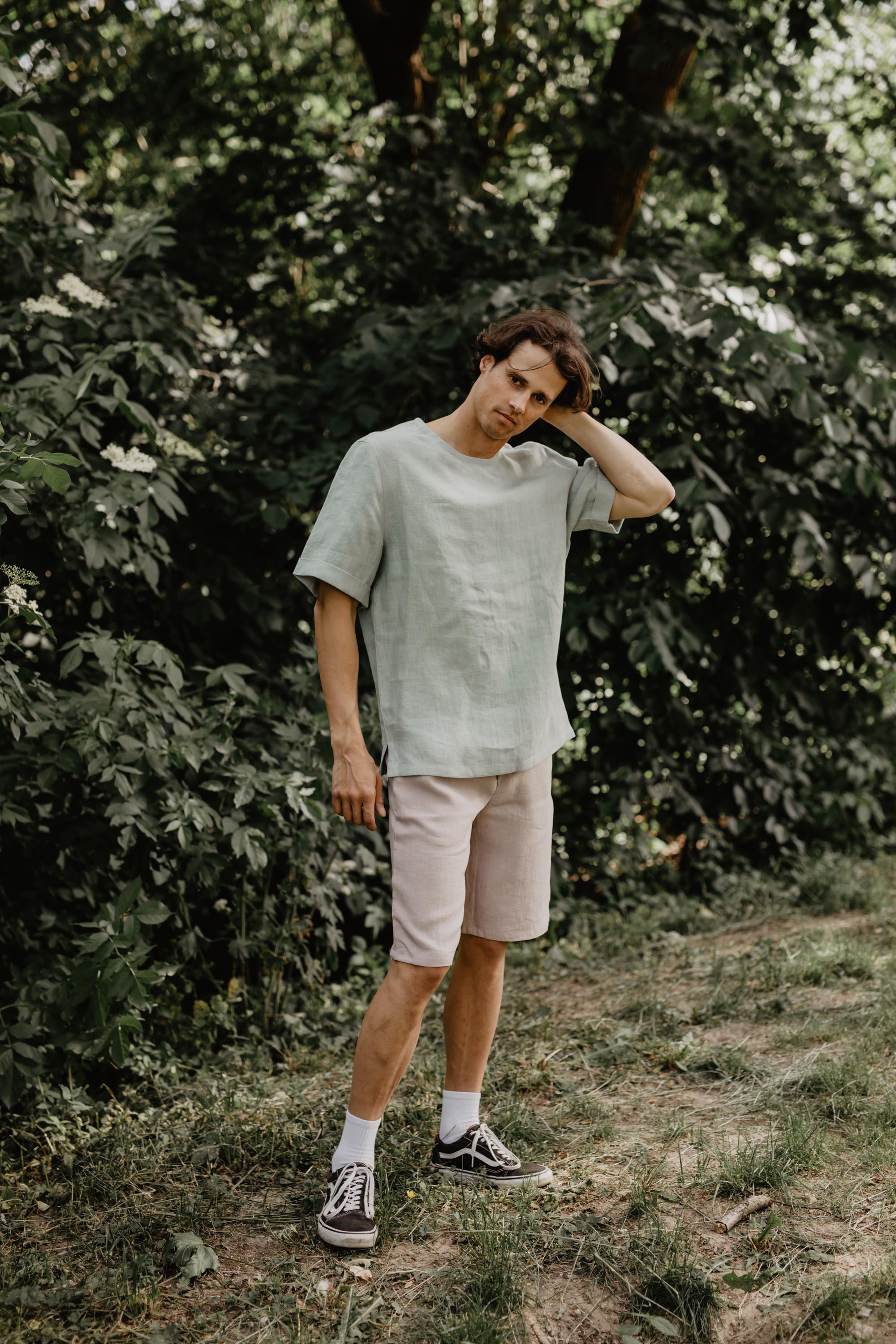 Man Facing Forward In Nature Wearing A Sage Green Linen Top And Light Linen Shorts