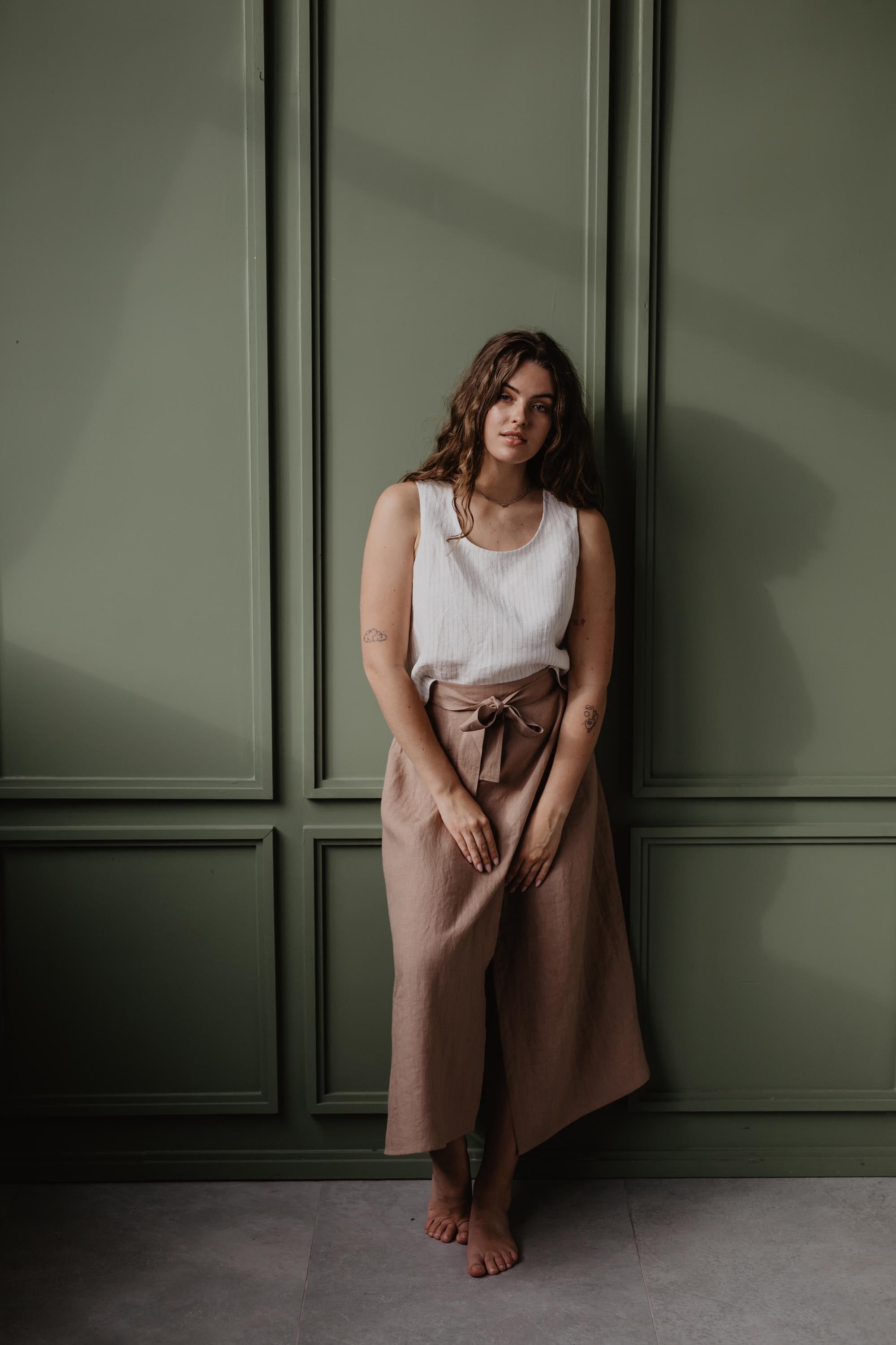 Woman Leaning On A Dark Green Wall Wearing White Pinafore Linen Top and A Dusty Rose Linen Long Skirt