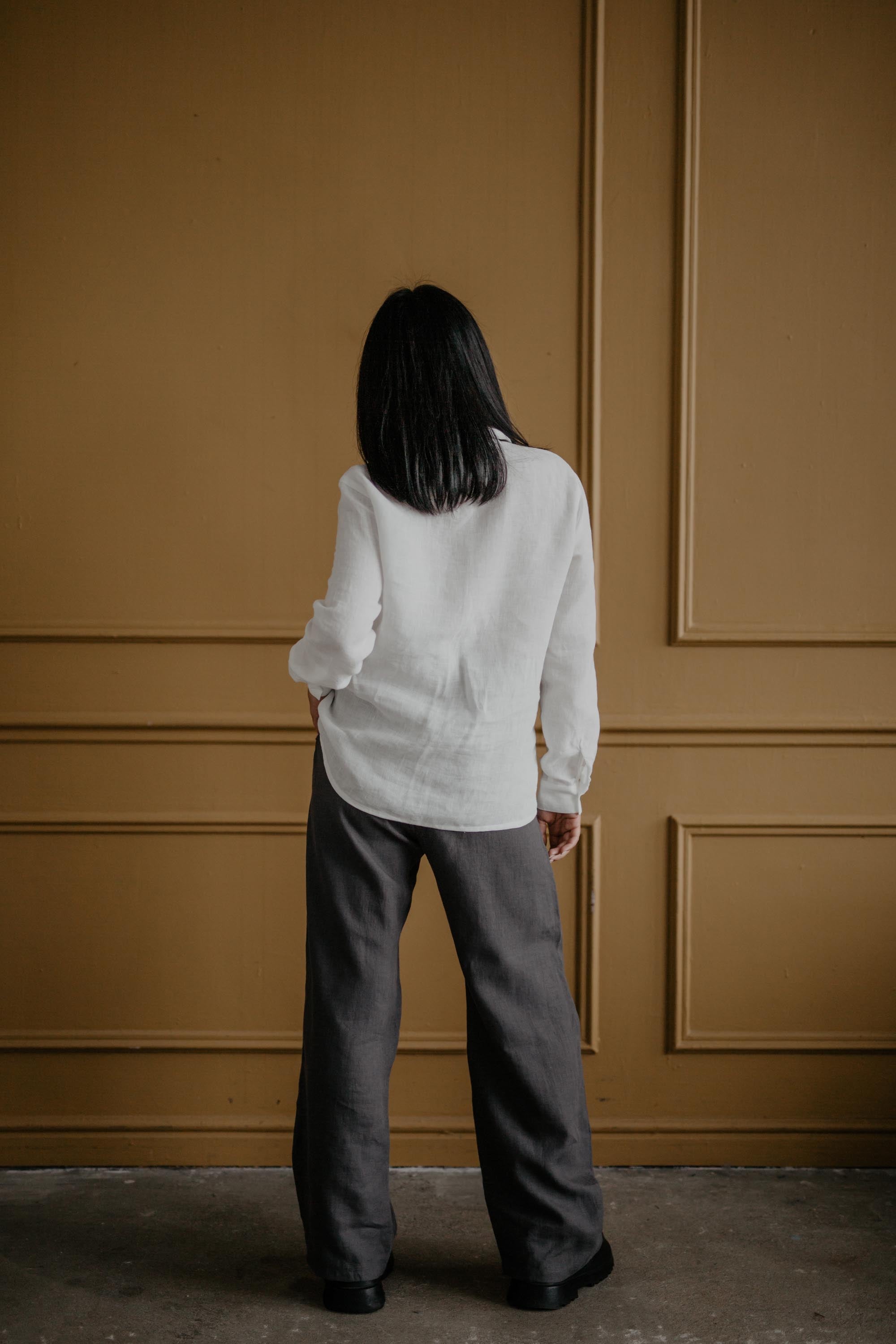 Woman Facing Back Wearing Super Long Grey Linen Pants And White Shirt In A Bronze Color Room