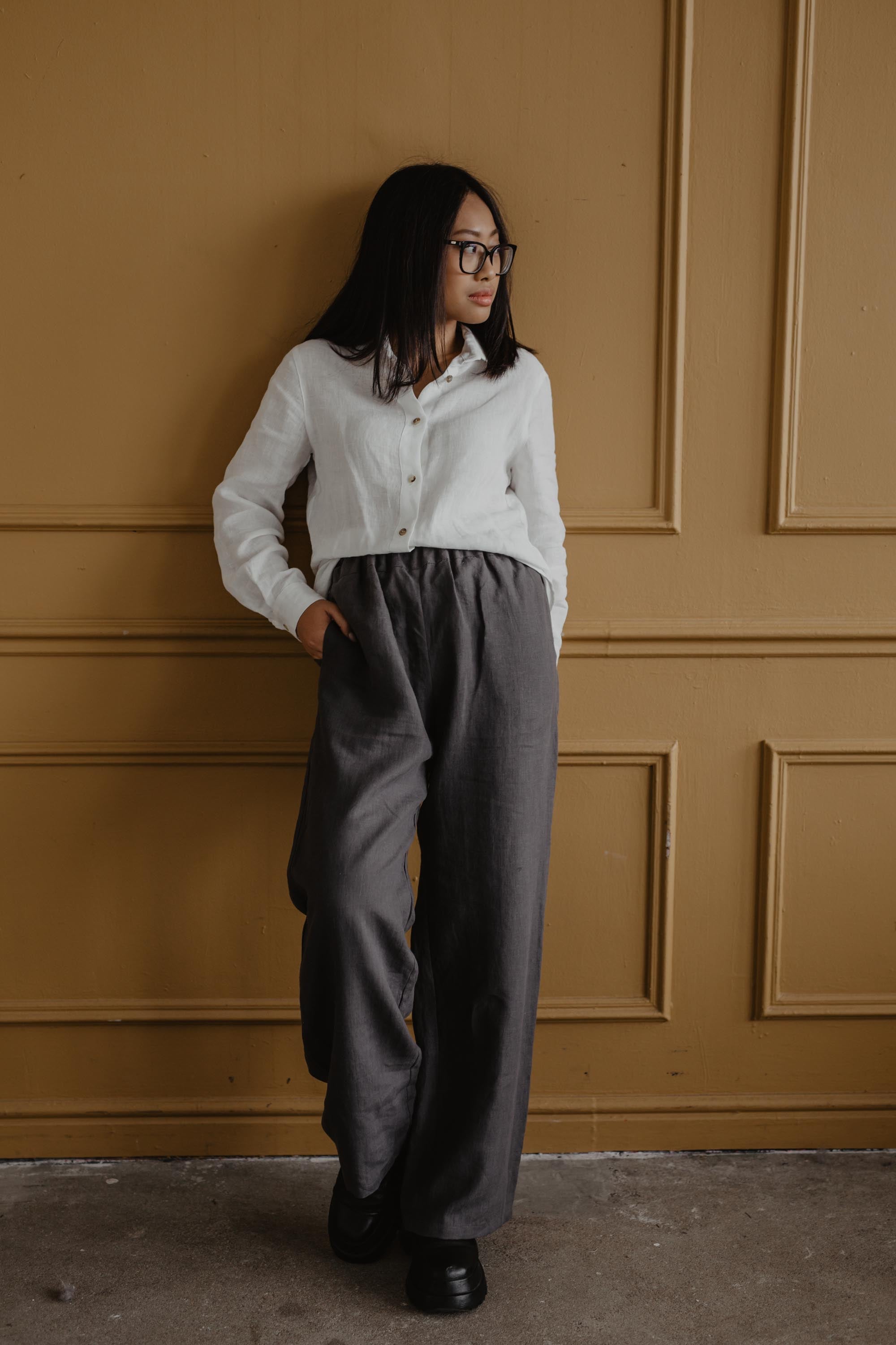 Woman Leaning Agains Wall Wearing Super Long Grey Linen Pants And White Linen Shirt