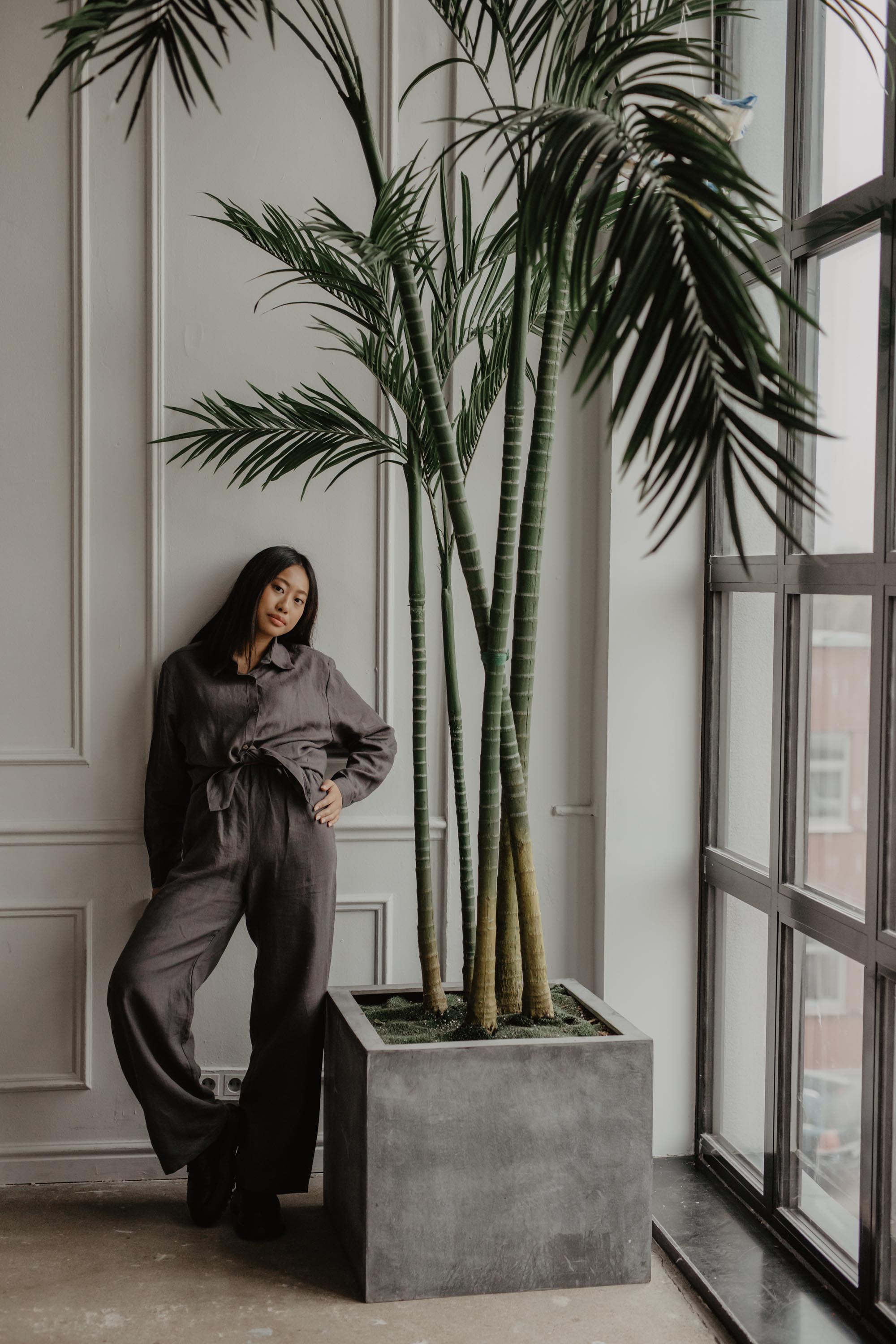 Woman Standing Next to Plant Wearing A Grey Casual Linen Shirt And Dark Linen Pants By AmourLinen