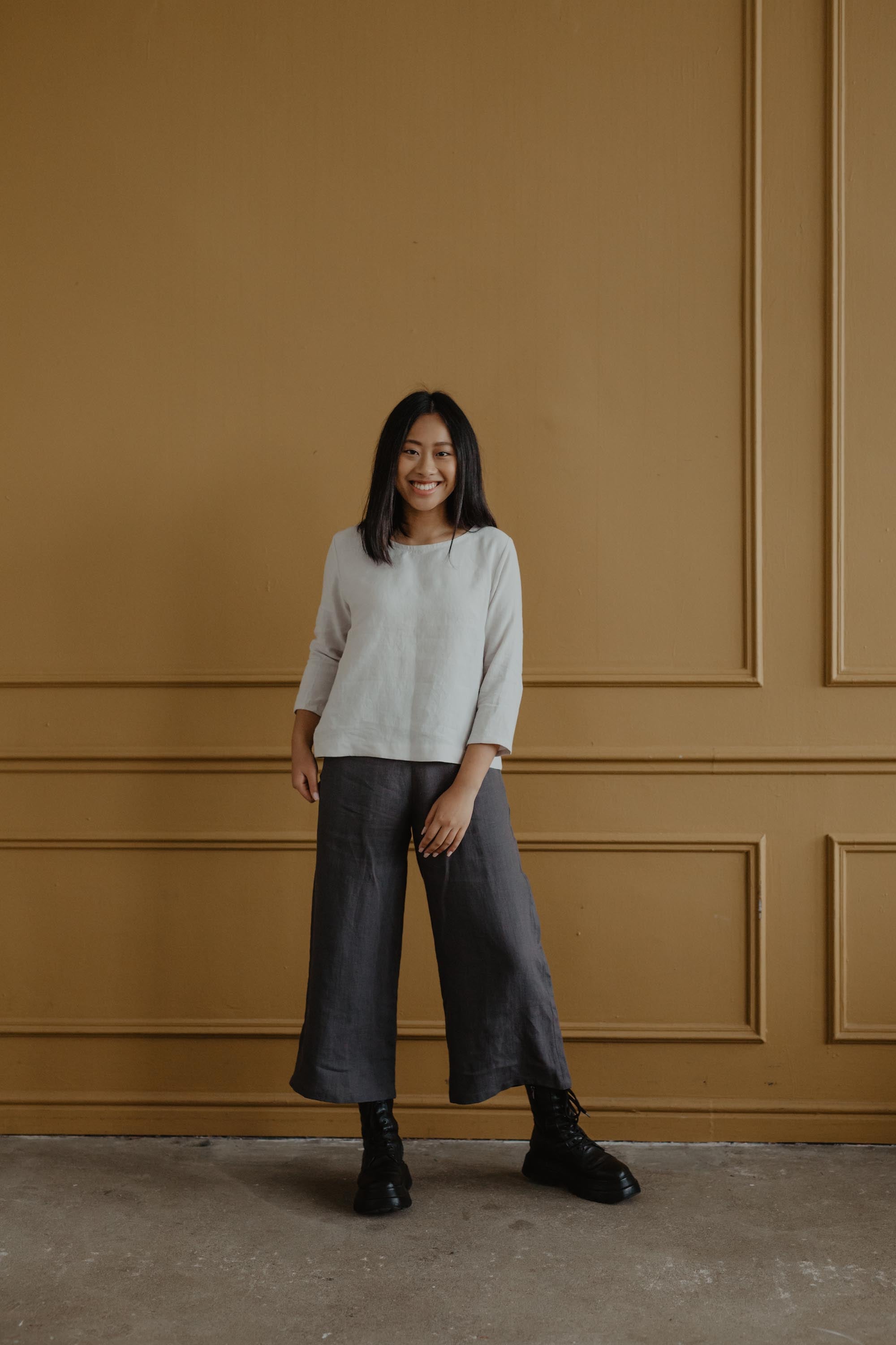 Woman Standing Next To A Bronze Wall Wearing A White Linen Top And Grey Linen Pants By AmourLInen