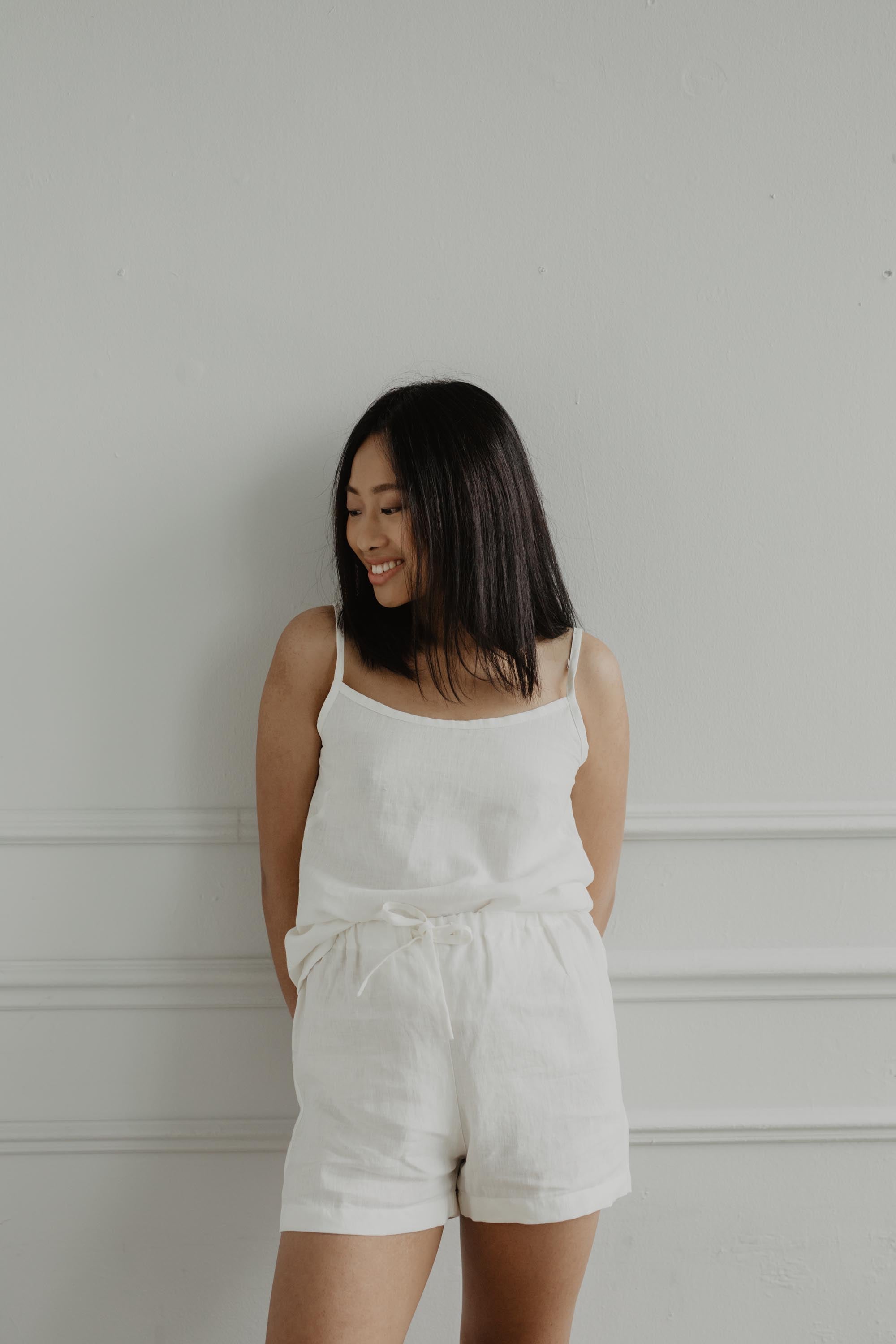 Woman Leaning On Wall Wearing A White Linen Sleeveless Pajama Set By AmourLinen