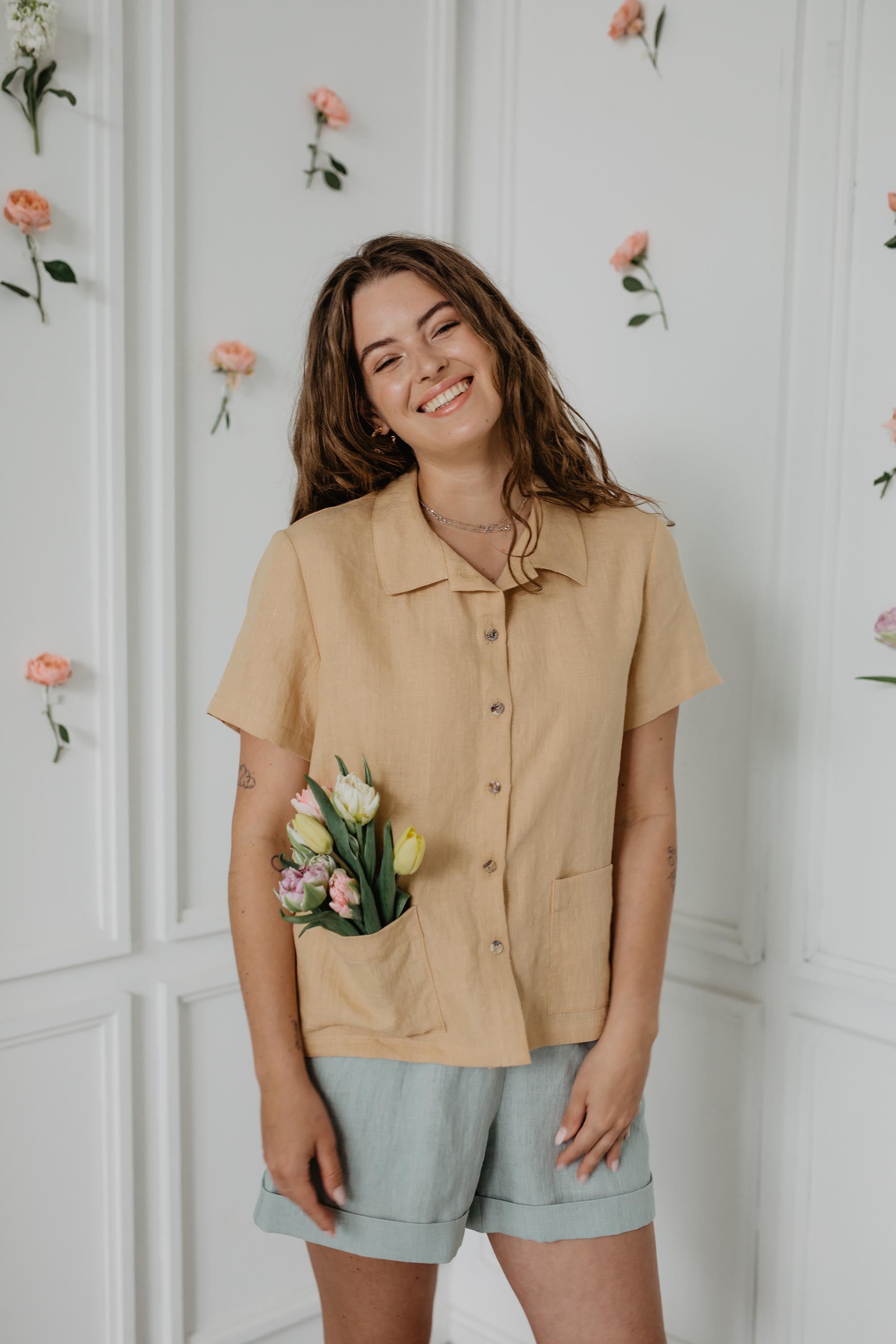 Woman Smiling In White Flowery Room Wearing A Mustard Color Summer Shirt And Sage Green Linen Shorts