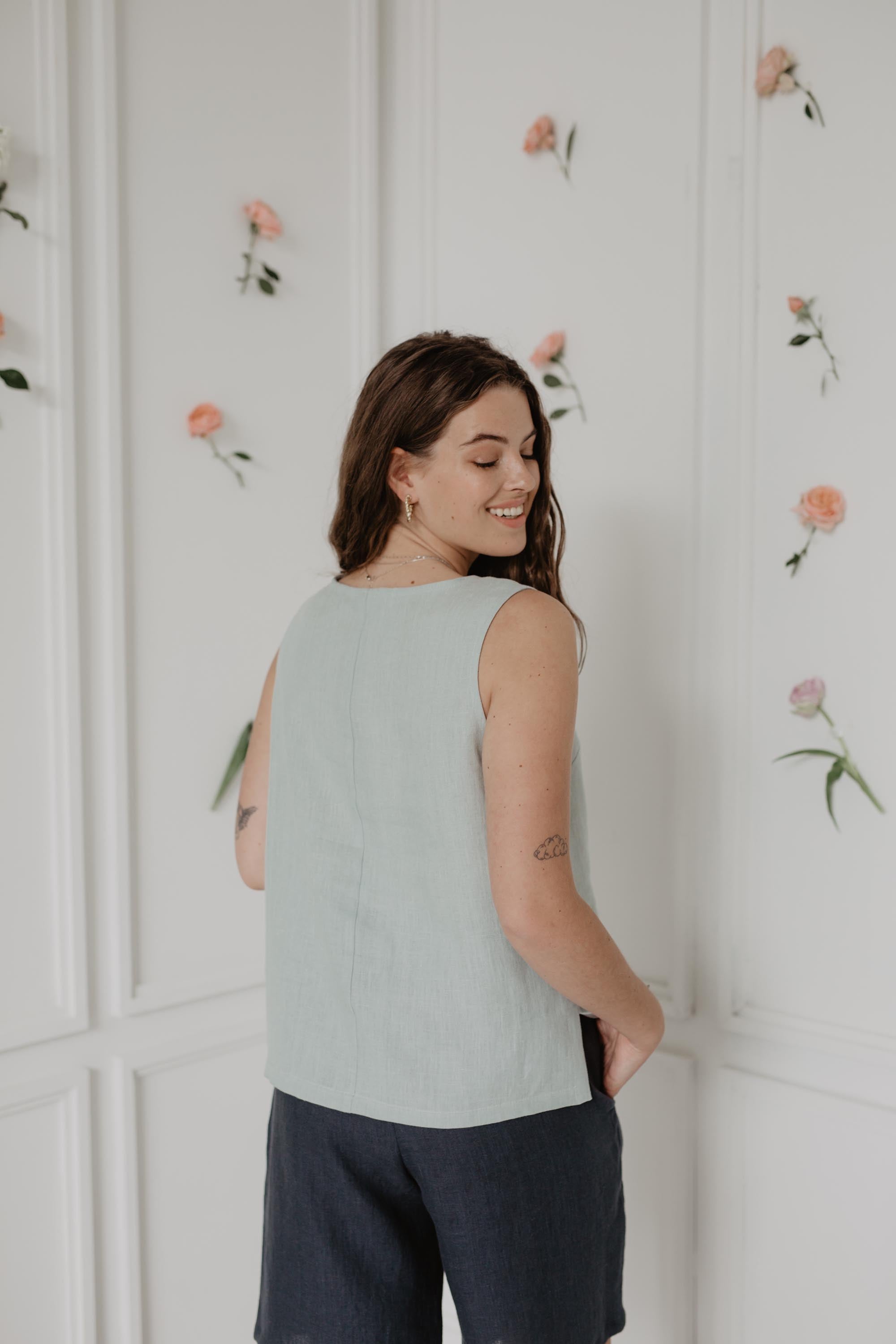 Back Of Woman Wearing A Sage Green Linen Classical Top and navy Blue Linen Shorts In Flowery Room