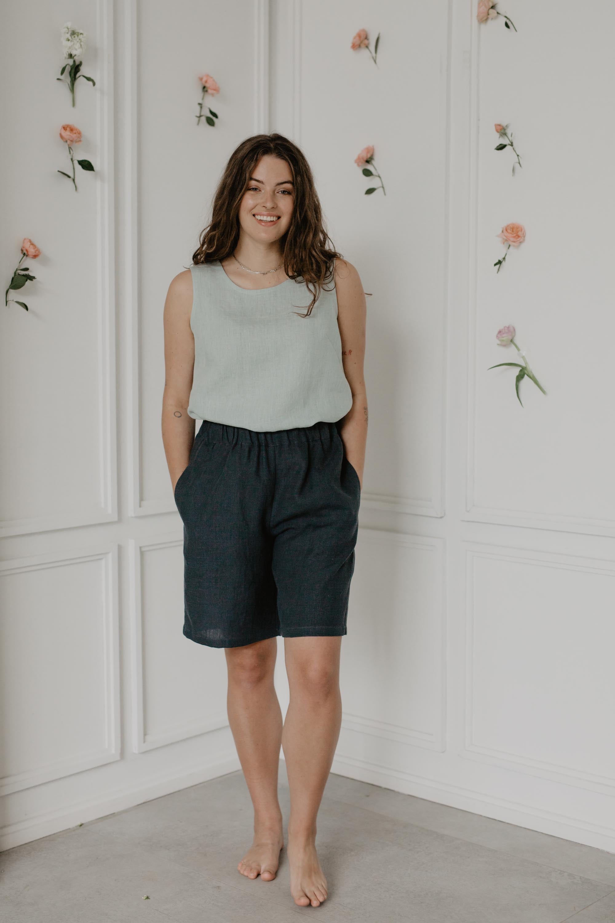 Woman In Flowery Room Wearing Charcoal Long Linen Shorts With A Sage Green Top