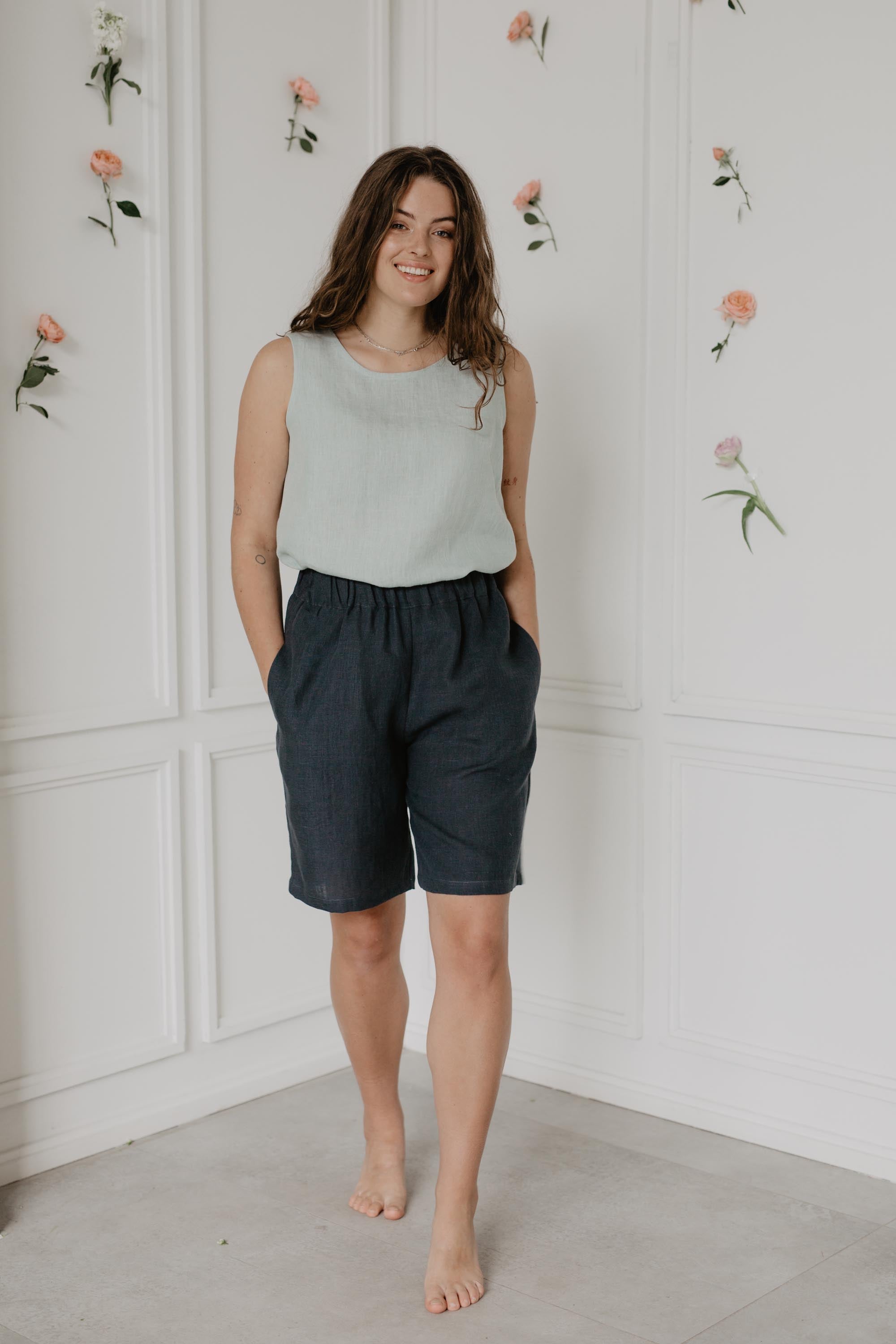 Woman Wearing Dark Long Linen Shorts And Sage Green Linen Top In A Light Room
