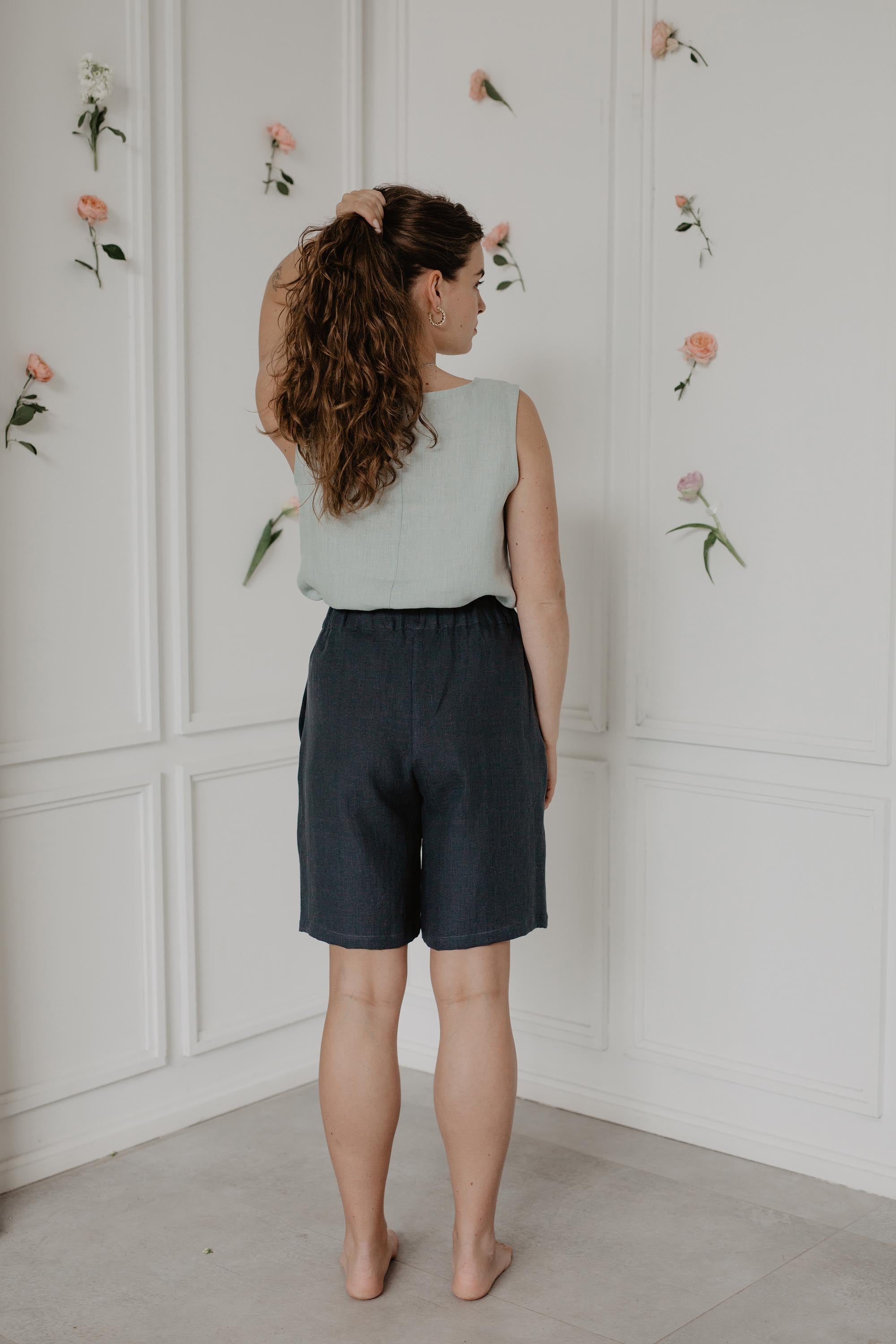 Back Of Woman Wearing Dark Linen Shorts And Sage Green Top In A Light Flowery Room