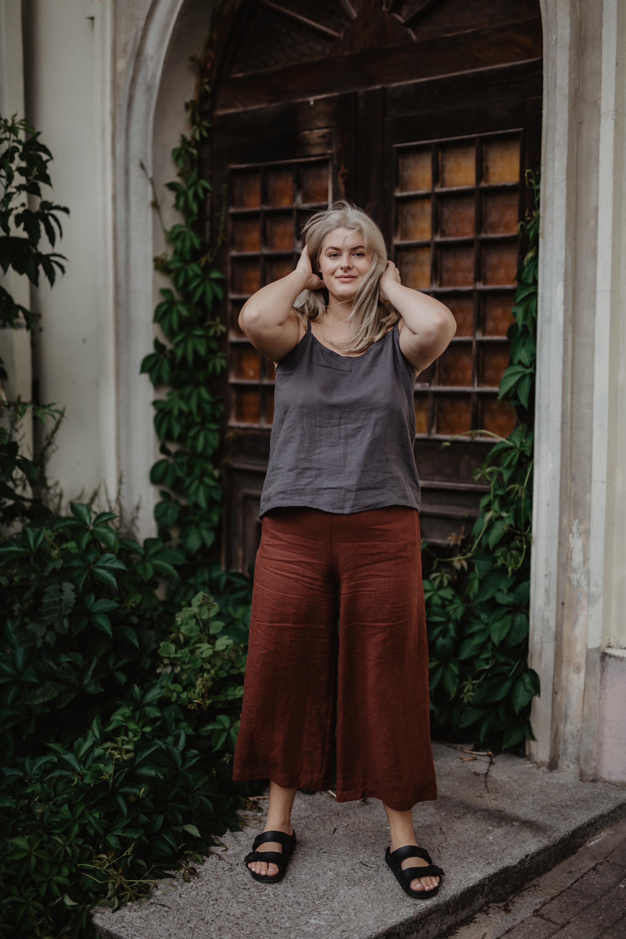 Woman Posing In Front Of a Botanical Door Wearing A Dark Linen Top And Teraccotta Linen Pants By Amourlinen