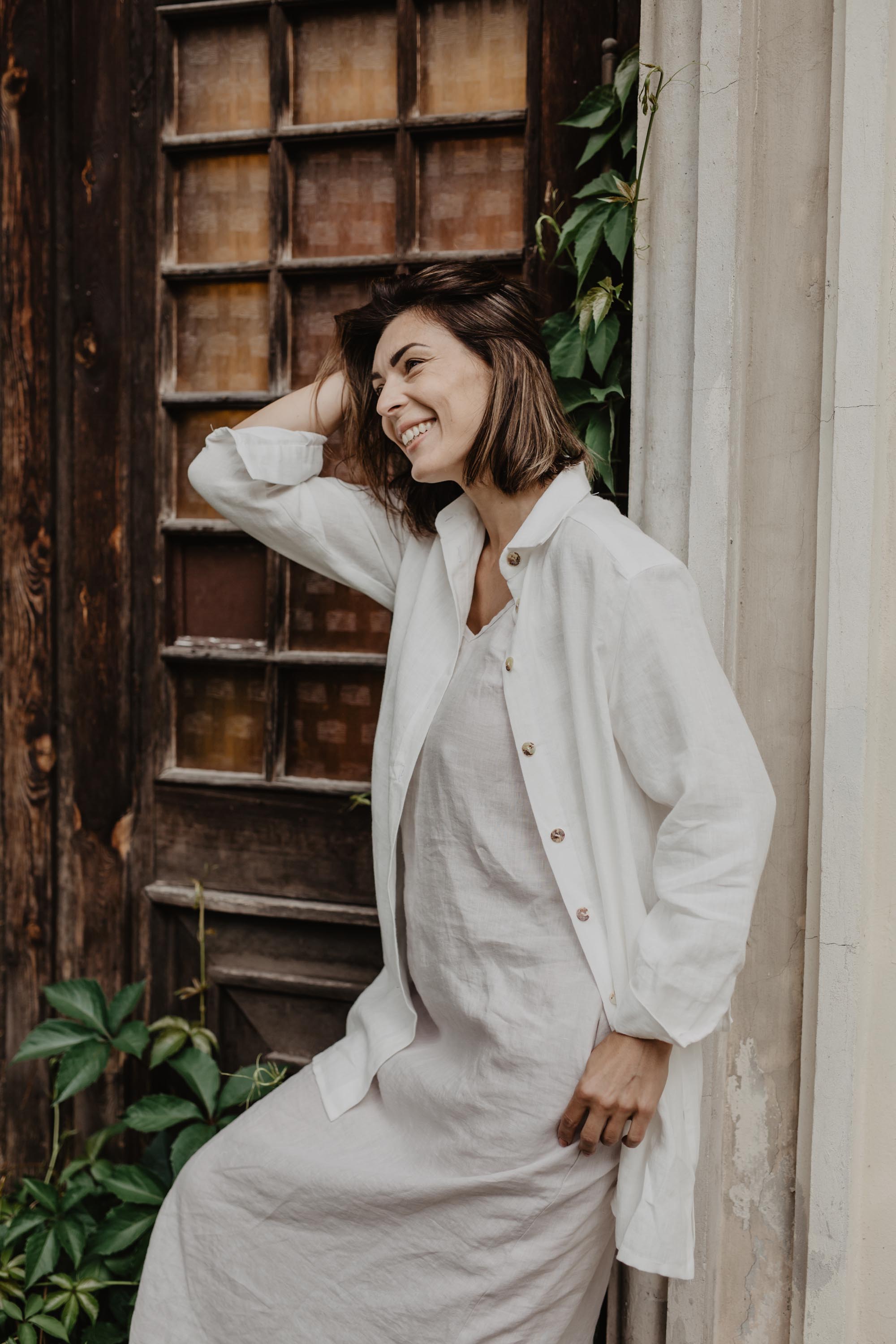 Woman Leaning Against A Wall Smiling Wearing A White Oversized Linen Shirt BY Amour Linen