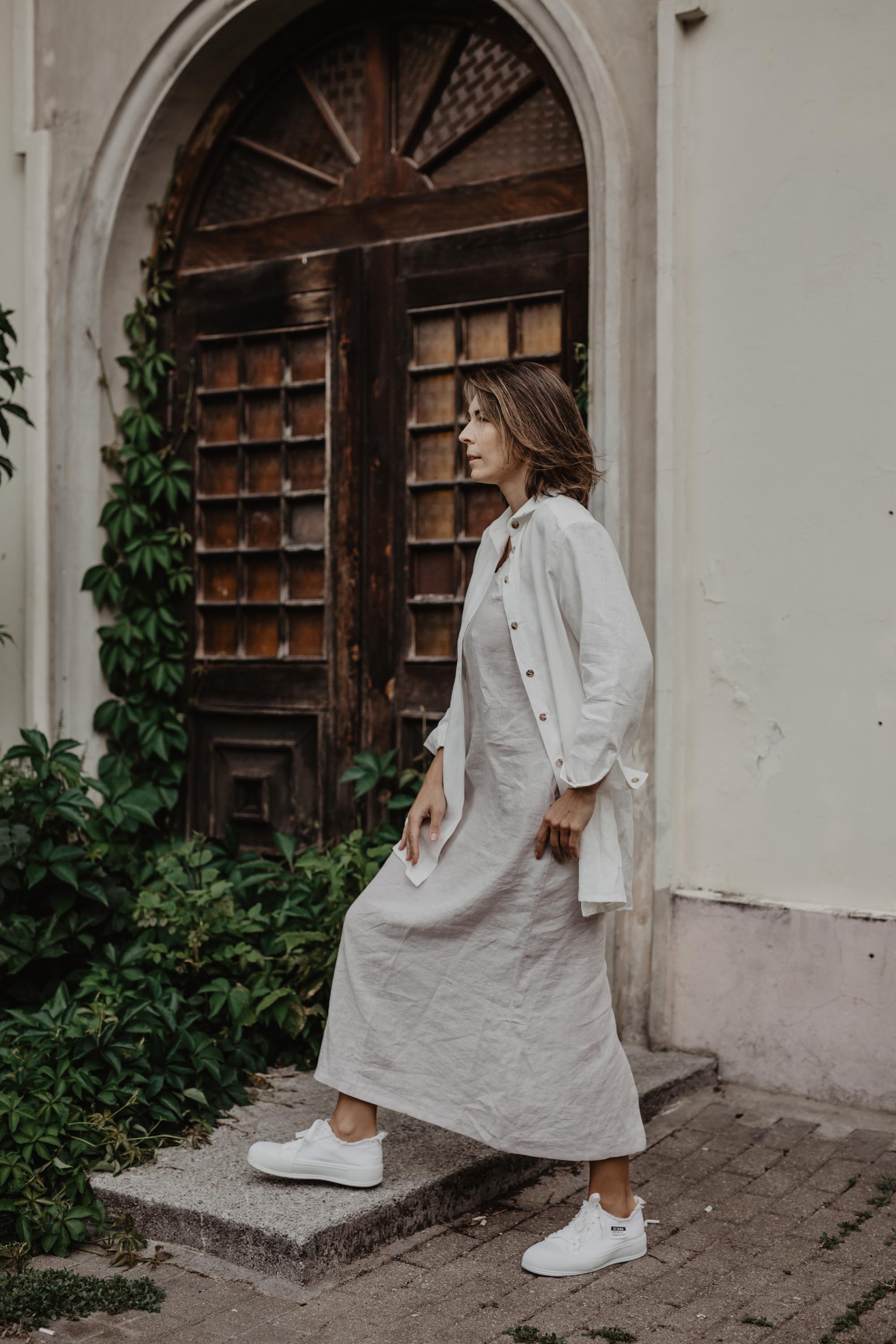 Woman Walking Next To A Botanical Door Wearing A White oversized Linen Shirt and White Linen Dress By Amourlinen