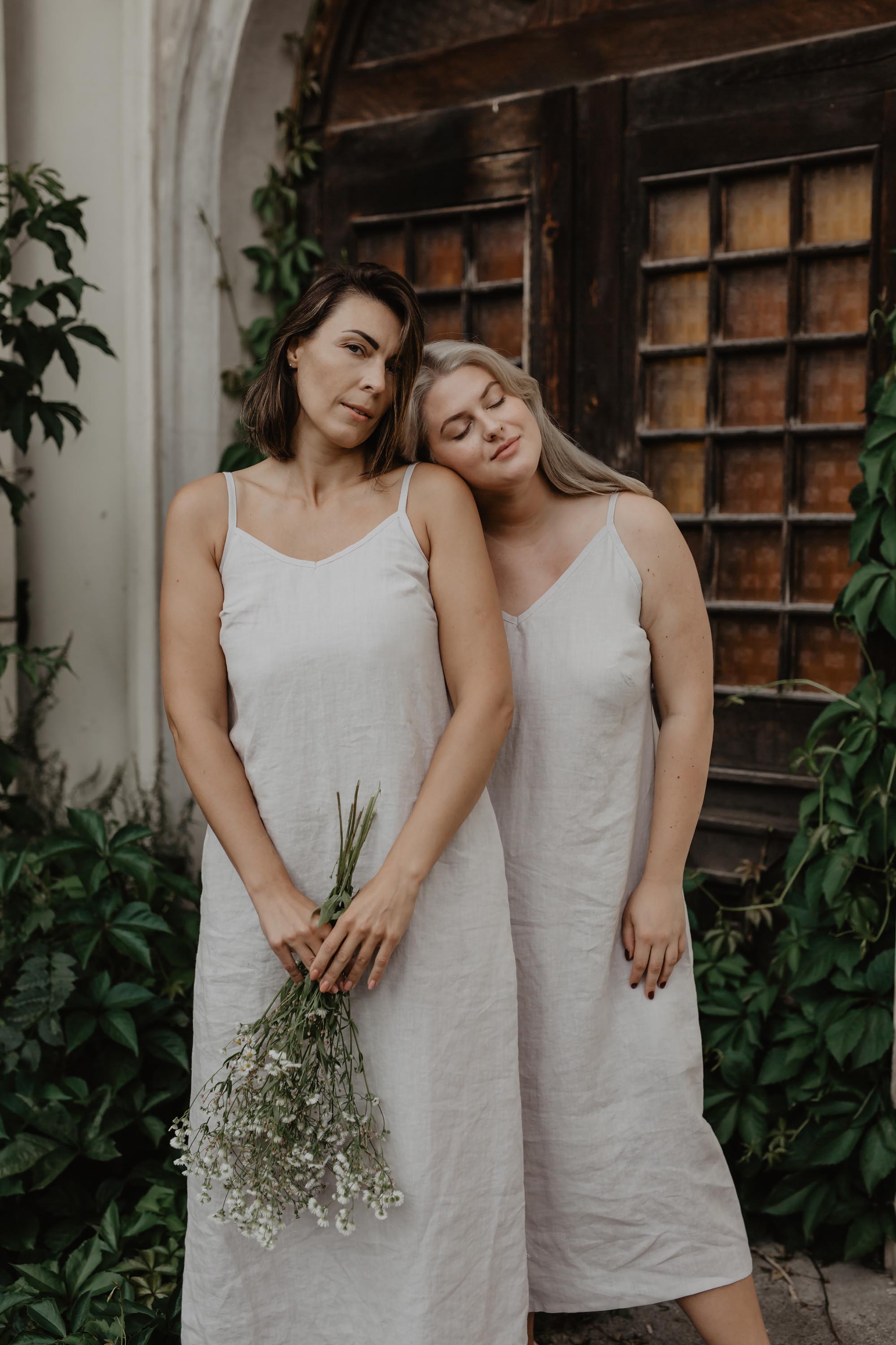 Two Women Wearing White Long Linen Desses BY AmourLinen Near A Botanical Door