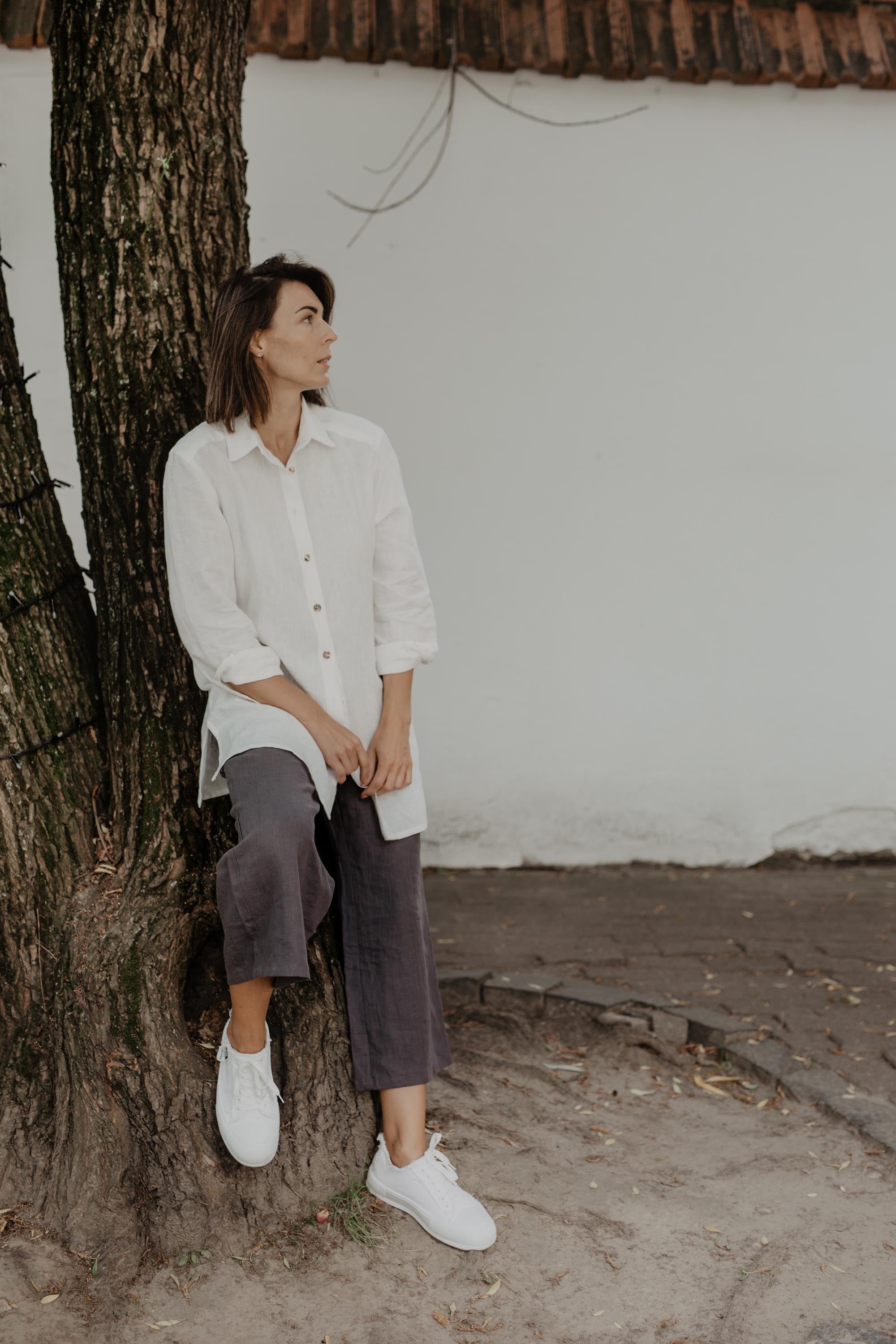 Woman Leanign Against A Tree Wearing A white Oversized Linen Shirt and Grey Linen Pants By Amourlinen