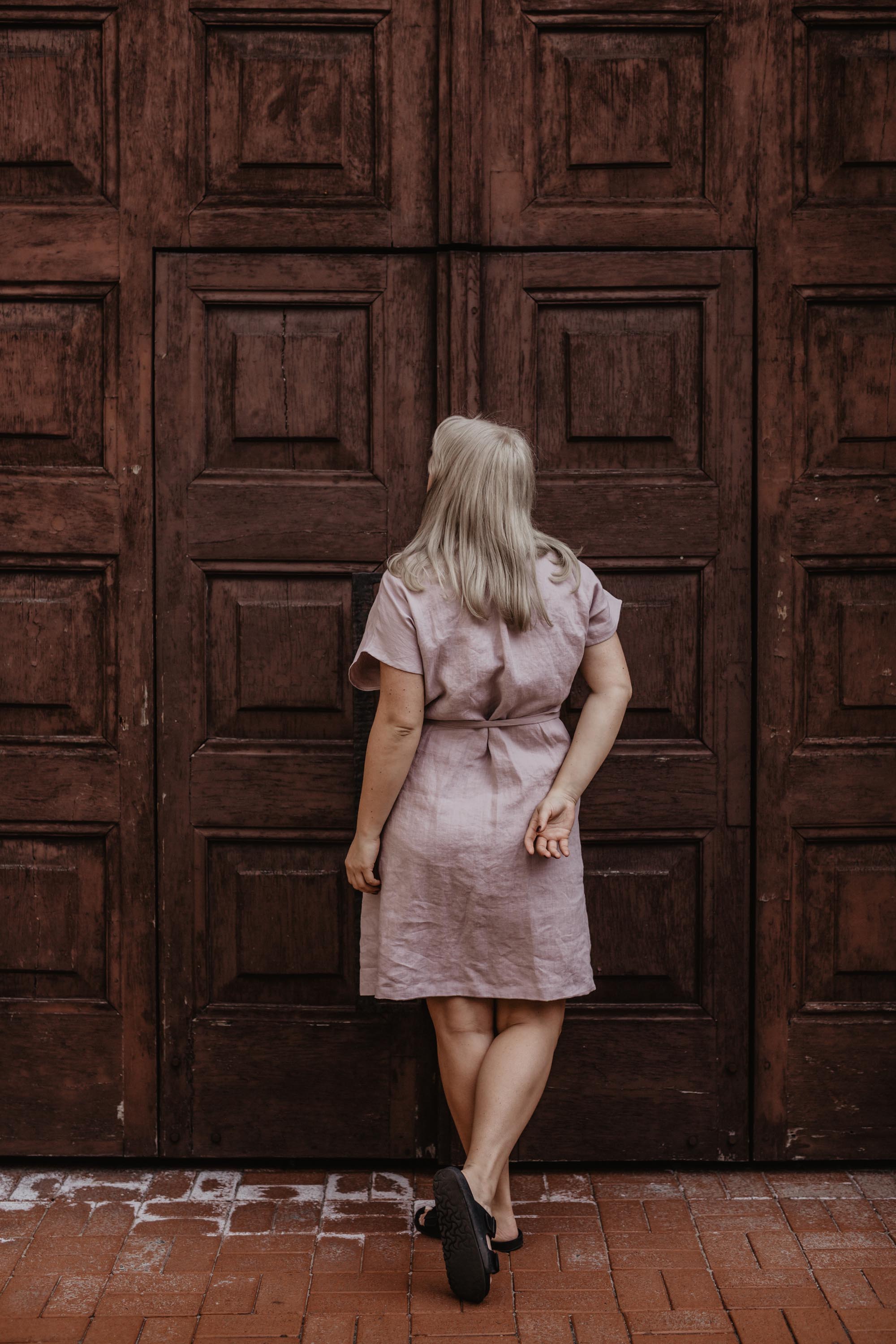 Woman Facing Back Looking At Huge Door Wearing A Dusty Rose Linen Wrap Dress