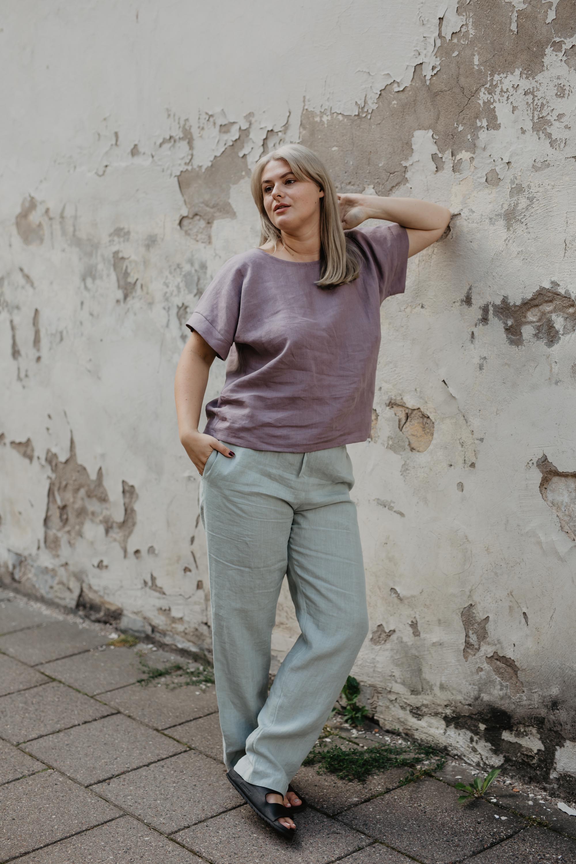 Woman Leaning Against A Wall Wearing Sage Green Linen Pants and Dusty Rose Linen Shirt By Amourlinen