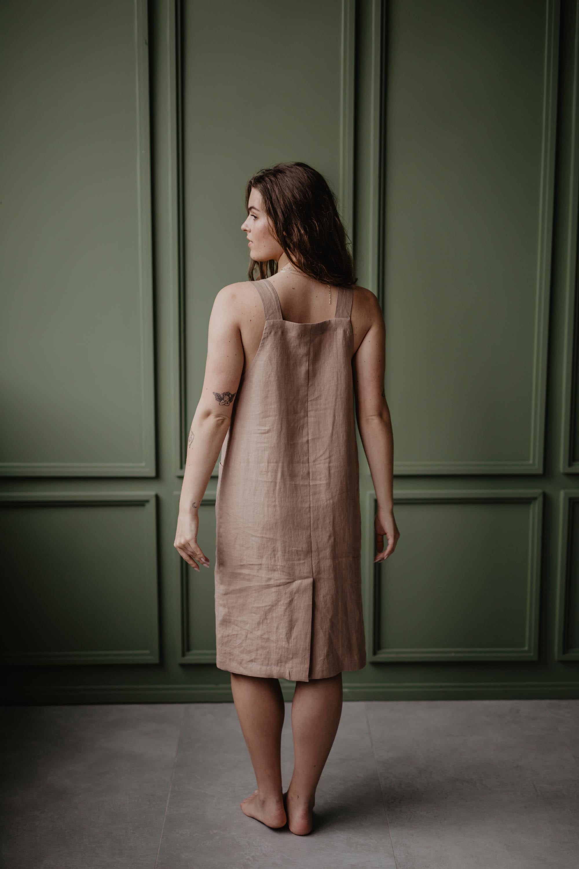 Back View Of A Women Wearing A Dusty Rose Apron Dress In A Green Room